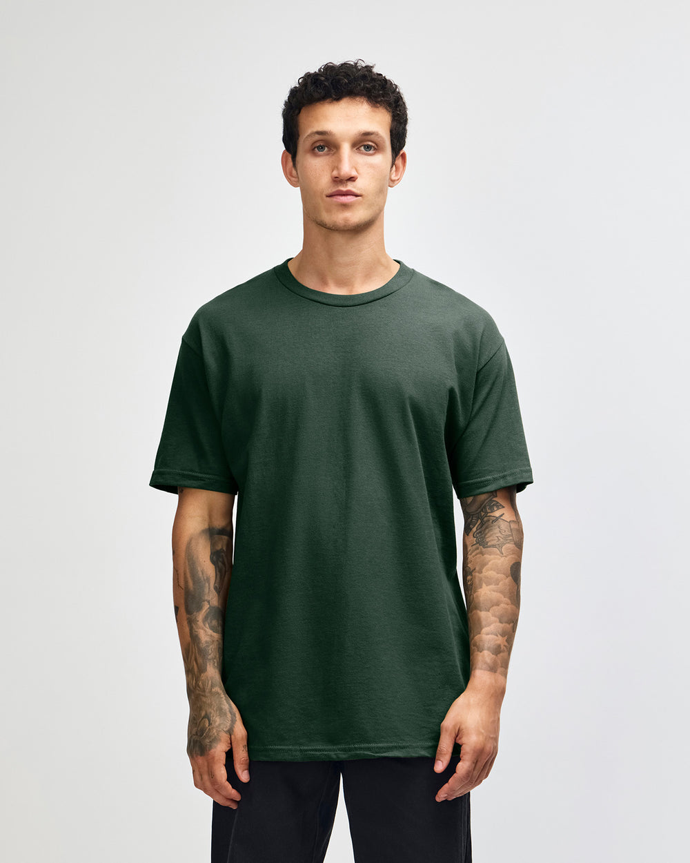 next image of Unisex Heavy T-shirt: Male Model wearing a Forest Heavyweight Unisex Short Sleeve Crew Neck Tee, paired with blue jeans, standing against a plain background (front pose) -forest green