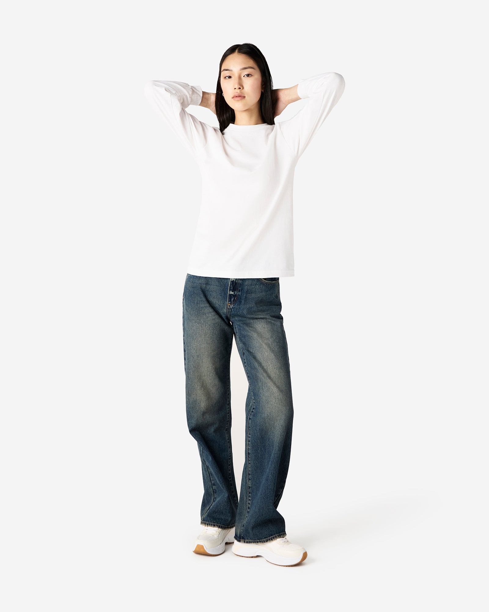 front view of a female Model wearing a White colored Heavyweight Unisex Long Sleeve Crew Neck Tee, paired with blue jeans, standing against a plain background (front pose) -white