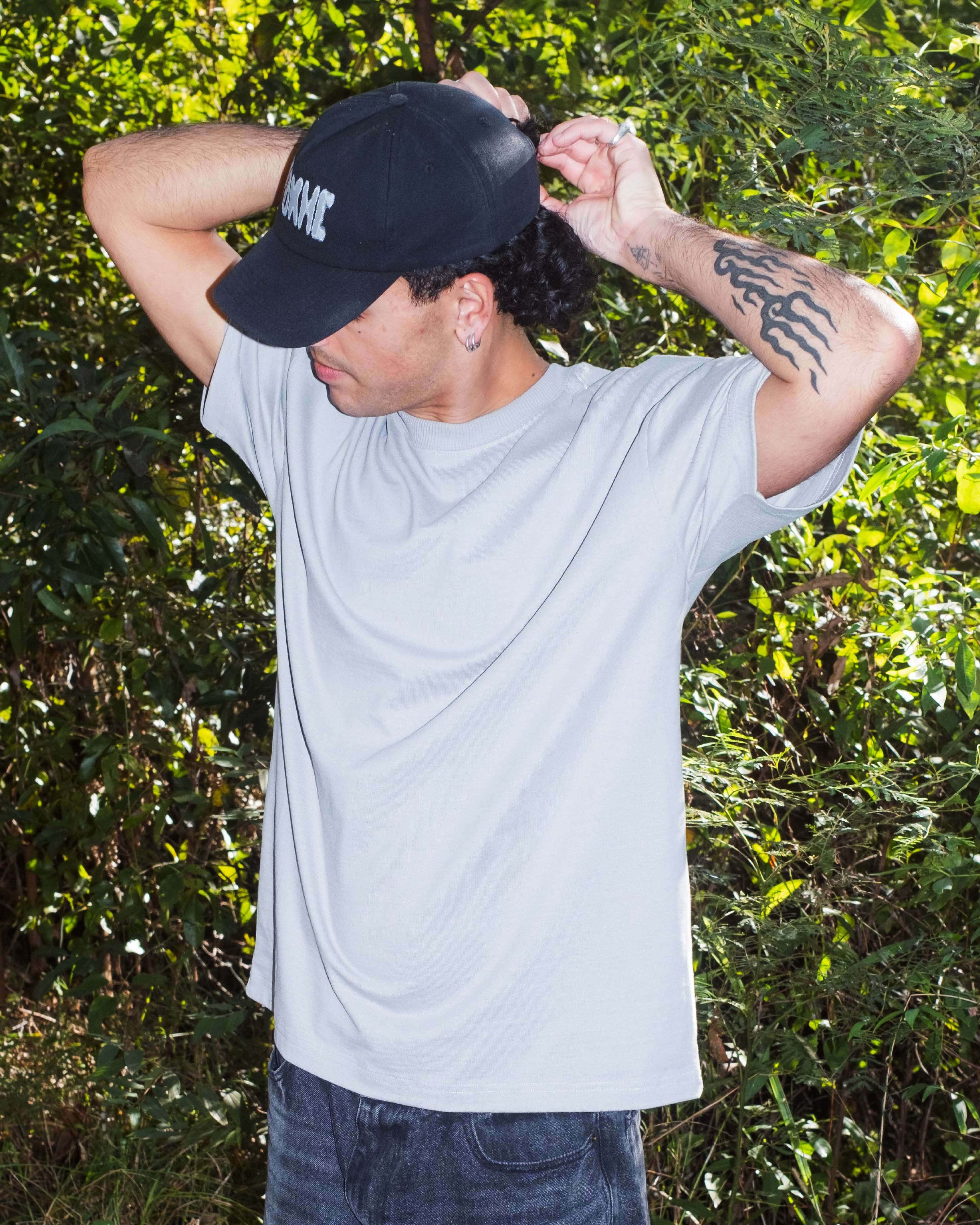 Person wearing a black cap and light gray t-shirt with a tattoo on arm, standing against a green leafy background