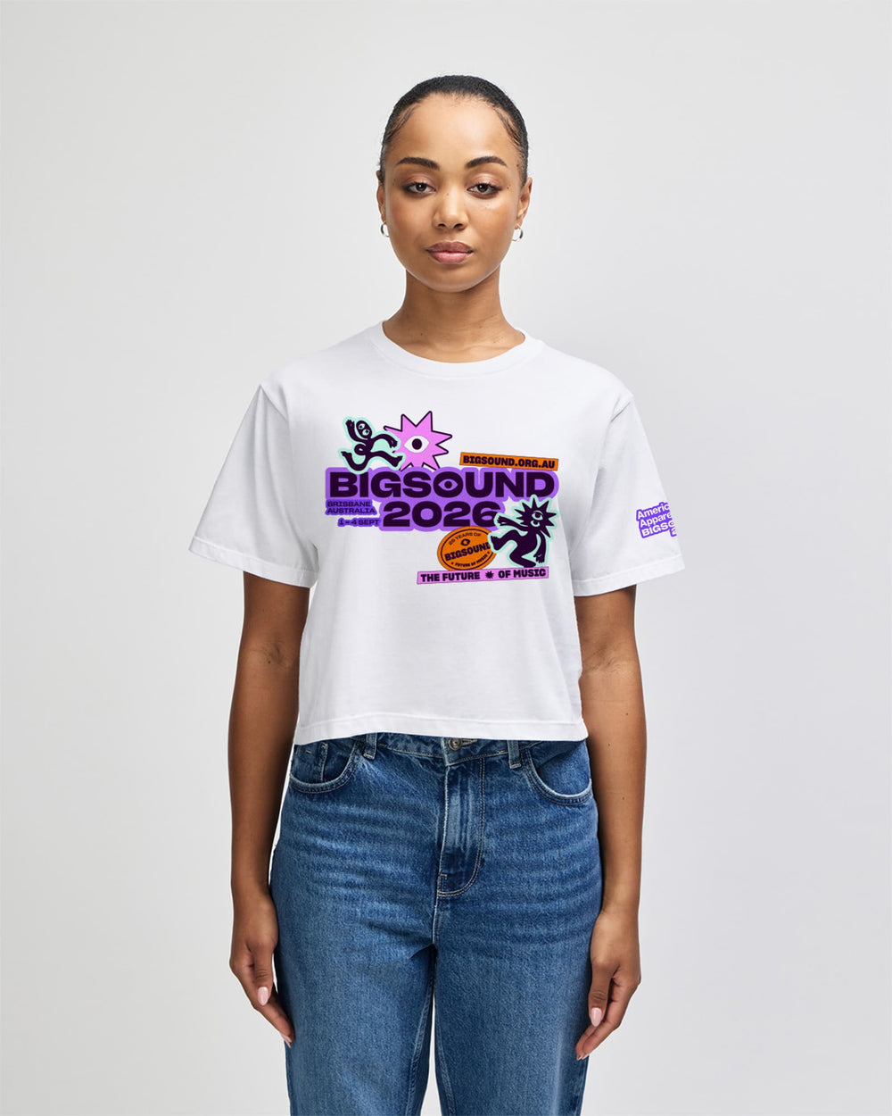 image of BIGSOUND Lineup Cropped T-shirt : A woman modeling a white cropped graphic T shirt with colorful 'BIGSOUND 2026' artwork on the front and a small purple graphic on the sleeve, paired with blue jeans against a light gray studio background -white