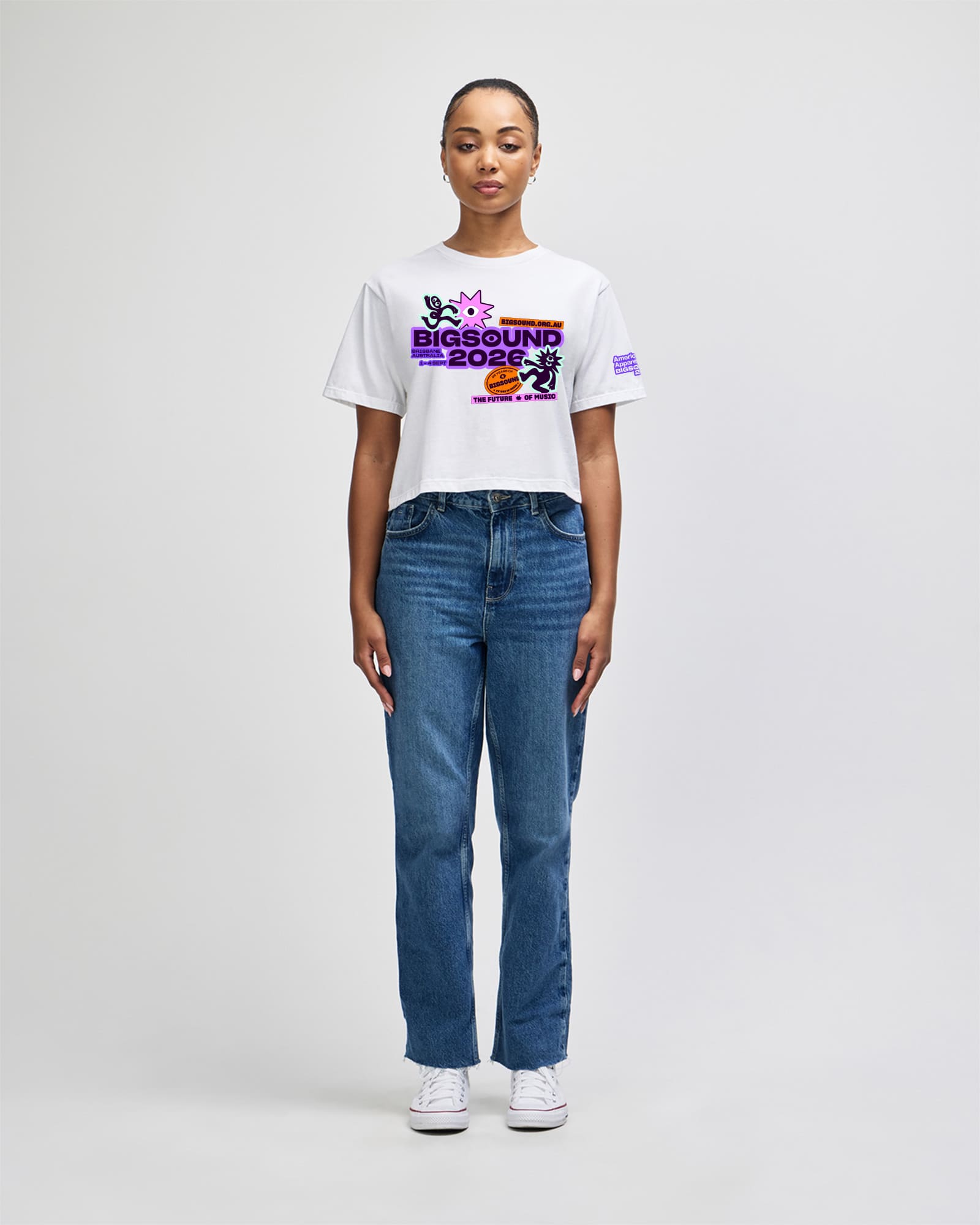 A model stands against a light gray studio background wearing a white graphic T shirt with a colorful 'BIG SOUND 2024' print on the chest, blue jeans, and white sneakers -white
