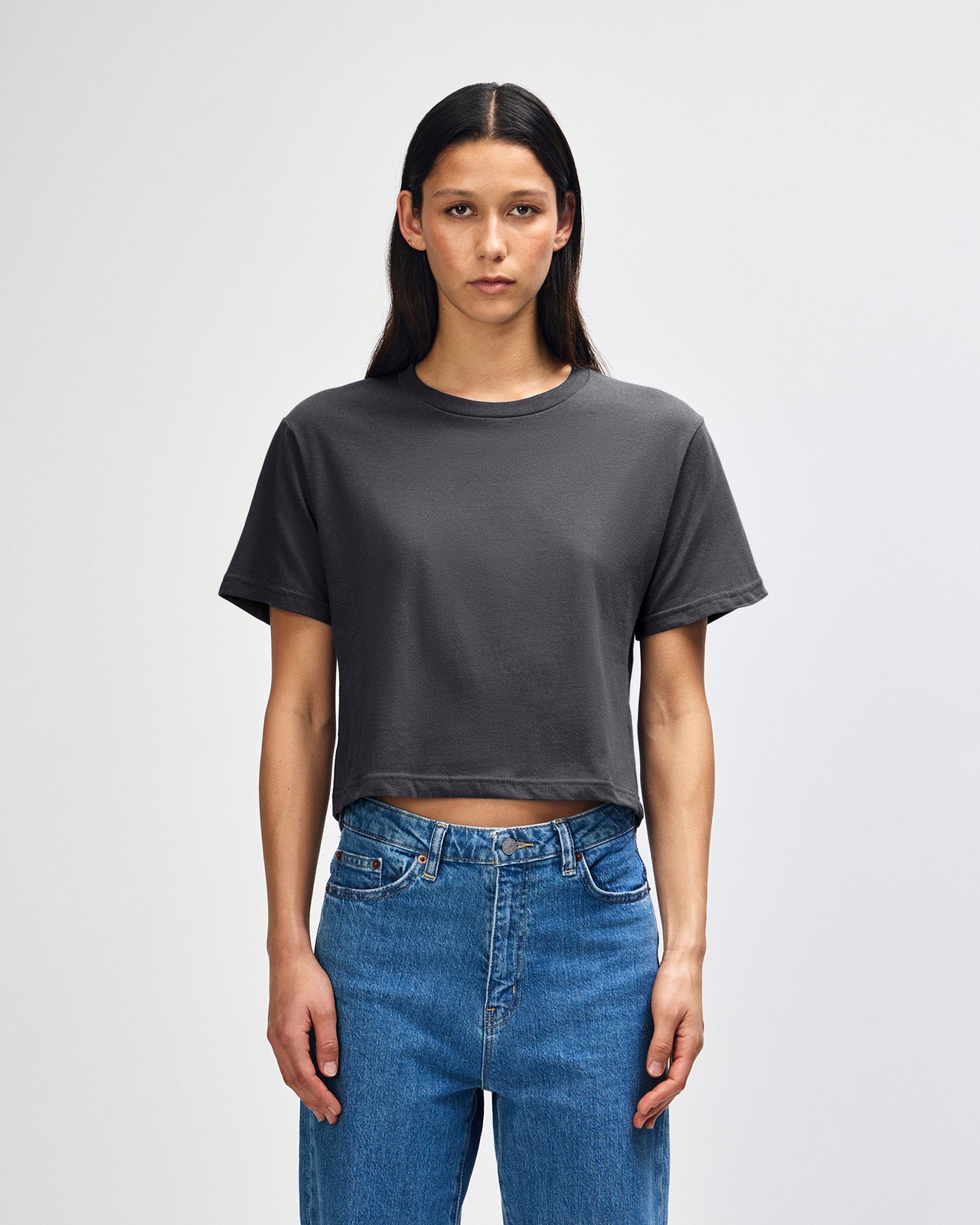 Female model wearing an American Apparel Fine Jersey Women's Boxy Tee in arctic, paired with blue jeans, standing against a plain background (front pose) -asphalt