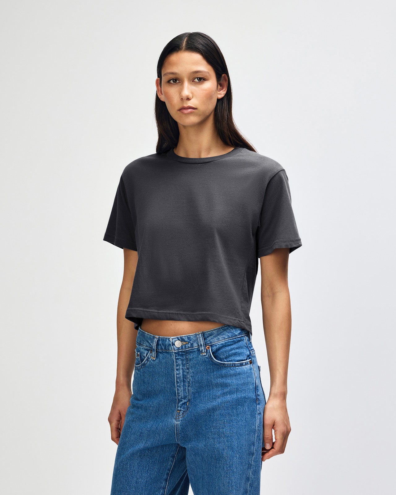 Female model wearing an American Apparel Fine Jersey Women's Boxy Tee in arctic, paired with blue jeans, standing against a plain background (front pose) -asphalt