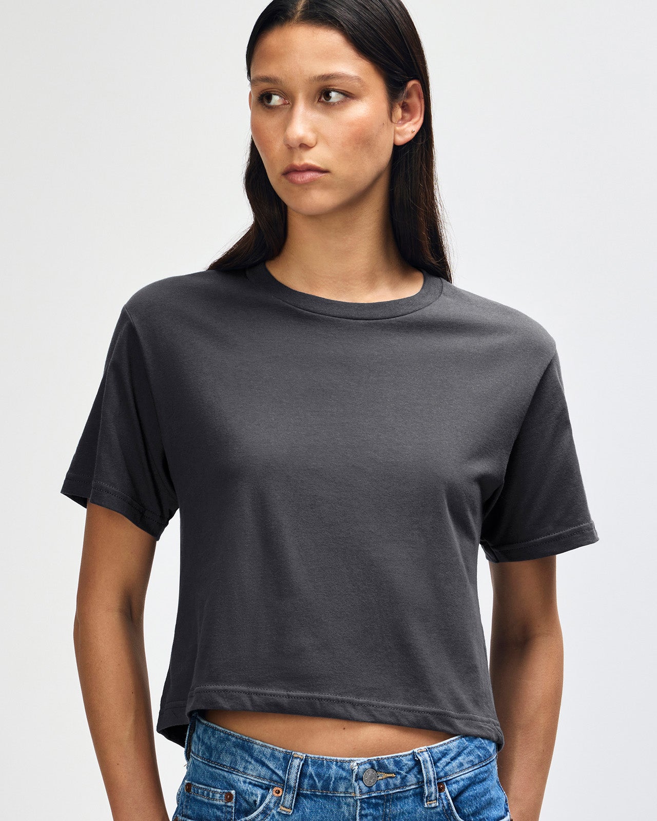 Female model wearing an American Apparel Fine Jersey Women's Boxy Tee in arctic, paired with blue jeans, standing against a plain background (front pose) -asphalt