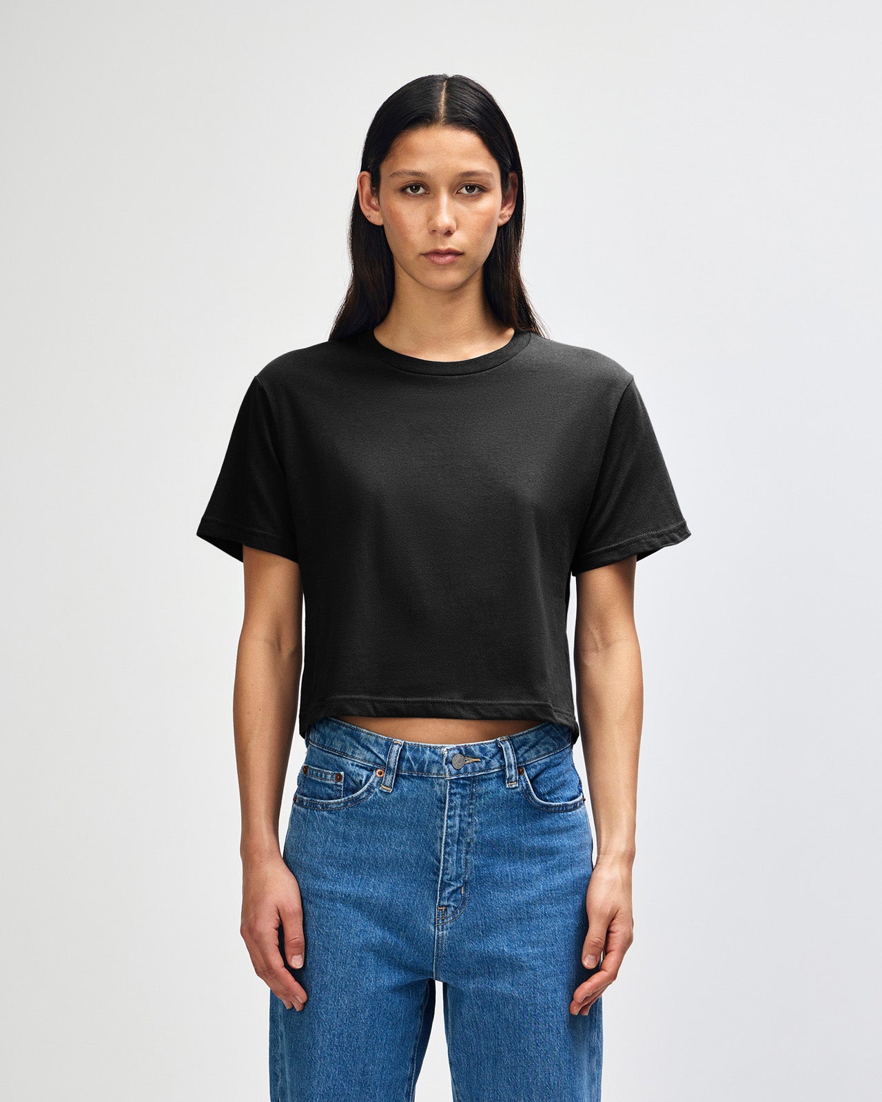 Female model wearing an American Apparel Fine Jersey Women's Boxy Tee in Black, paired with blue jeans, standing against a plain background (front pose) -black