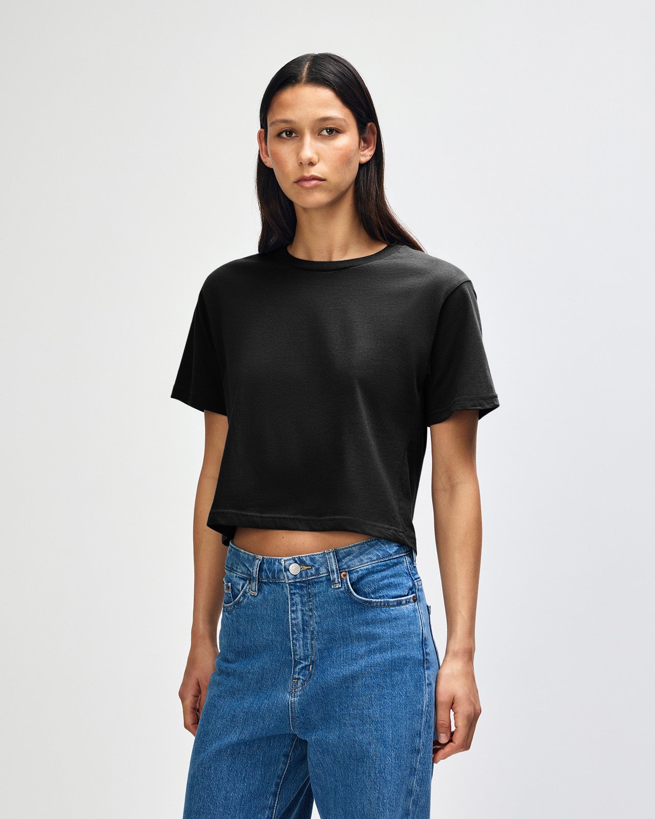 Female model wearing an American Apparel Fine Jersey Women's Boxy Tee in Black, paired with blue jeans, standing against a plain background (front pose) -black