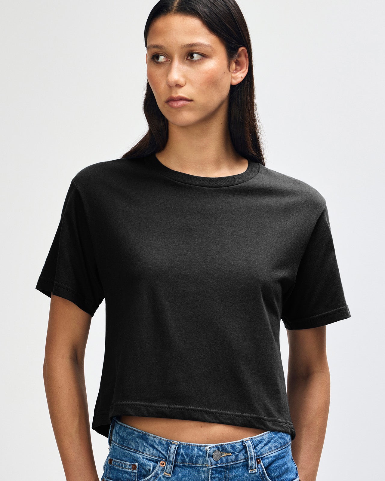 Female model wearing an American Apparel Fine Jersey Women's Boxy Tee in Black, paired with blue jeans, standing against a plain background (front pose) -black