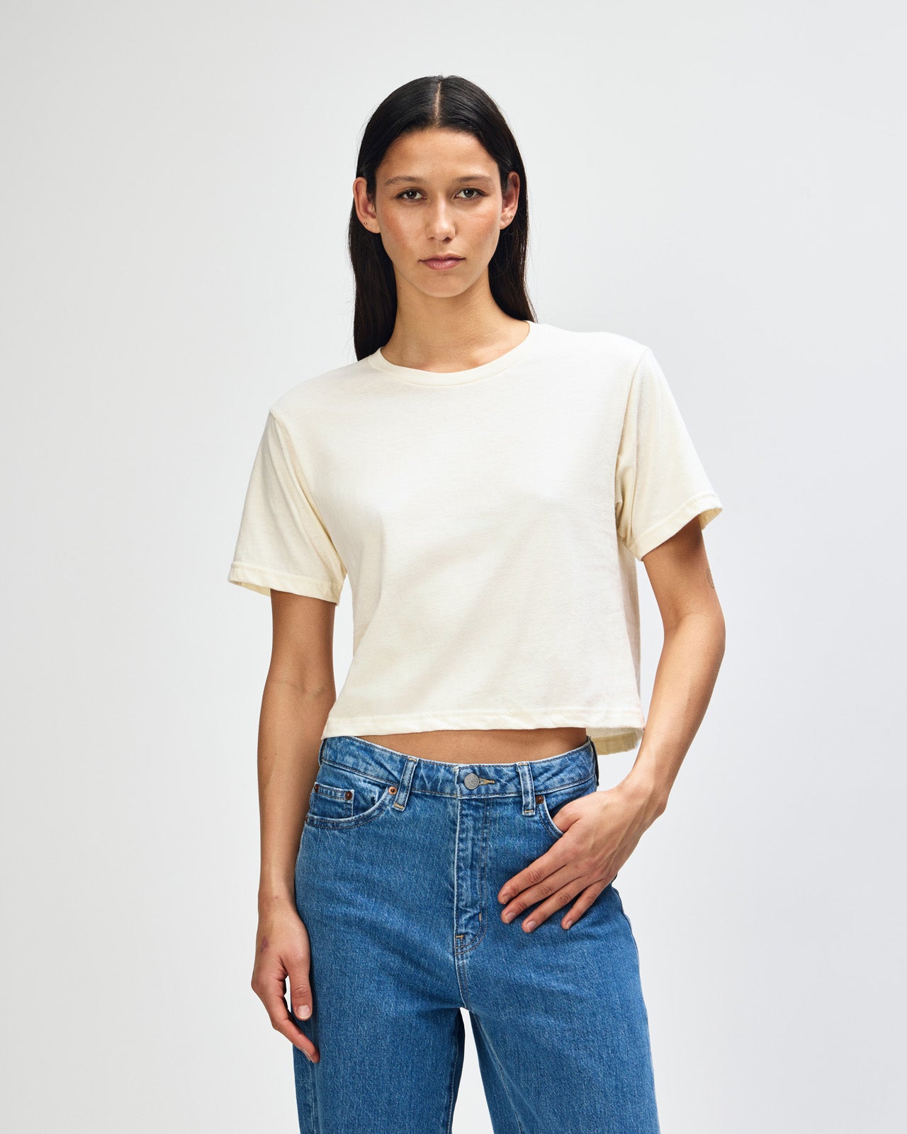 Female model wearing an American Apparel Fine Jersey Women's Boxy Tee in cream, paired with blue jeans, standing against a plain background (front pose) -cream