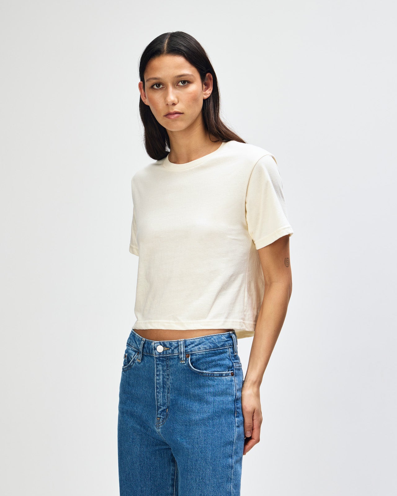 Female model wearing an American Apparel Fine Jersey Women's Boxy Tee in cream, paired with blue jeans, standing against a plain background (front pose) -cream
