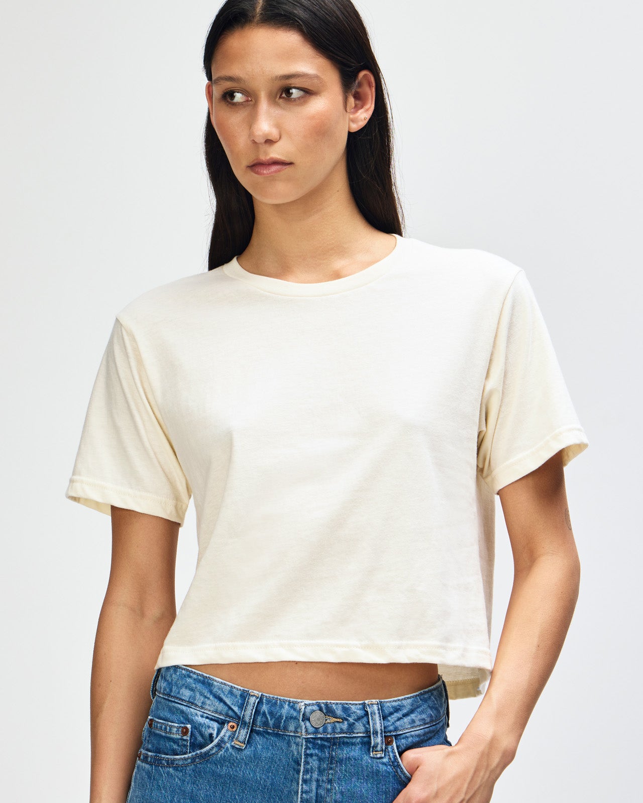 Female model wearing an American Apparel Fine Jersey Women's Boxy Tee in cream, paired with blue jeans, standing against a plain background (front pose) -cream