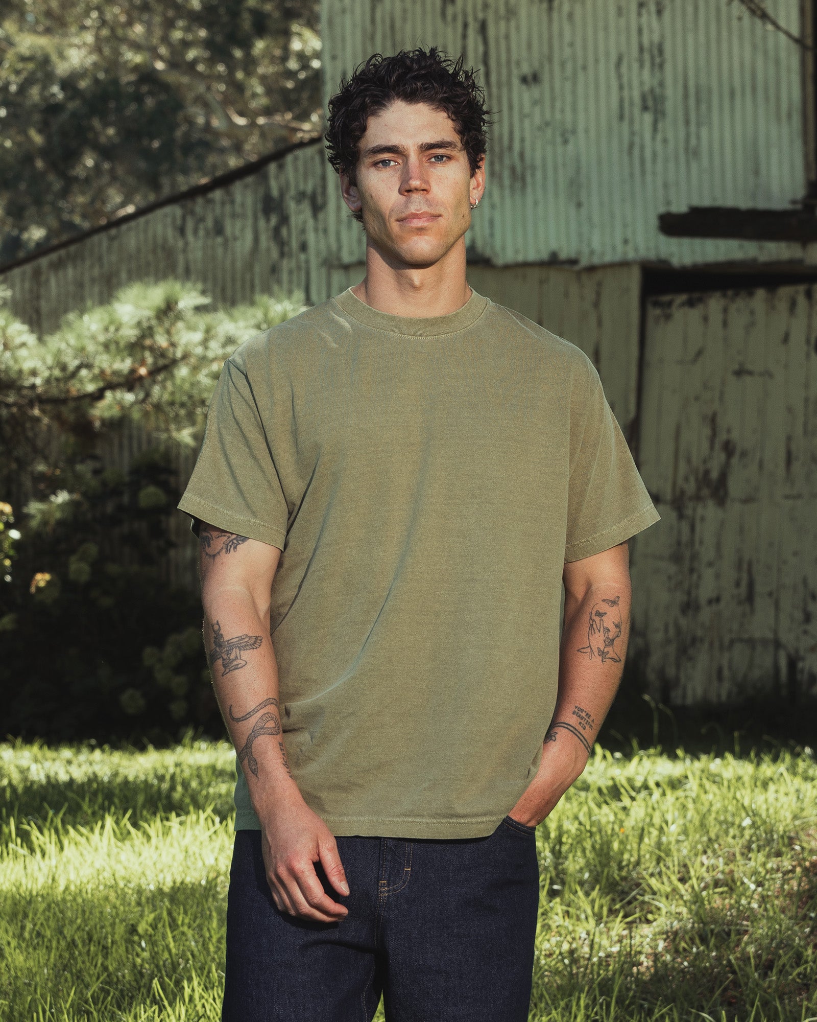 Male model wearing an American Apparel Garment Dyed Heavyweight Cotton Tee Shirt in Faded Army, paired with light blue jeans, standing against a plain background (front pose) -army
