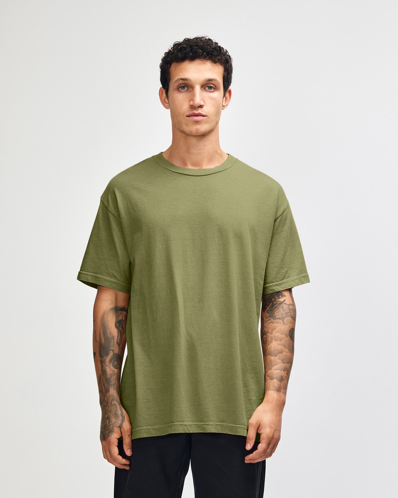 Male model wearing an American Apparel Garment Dyed Heavyweight Cotton Tee Shirt in Faded Army, paired with light blue jeans, standing against a plain background (front pose) -army