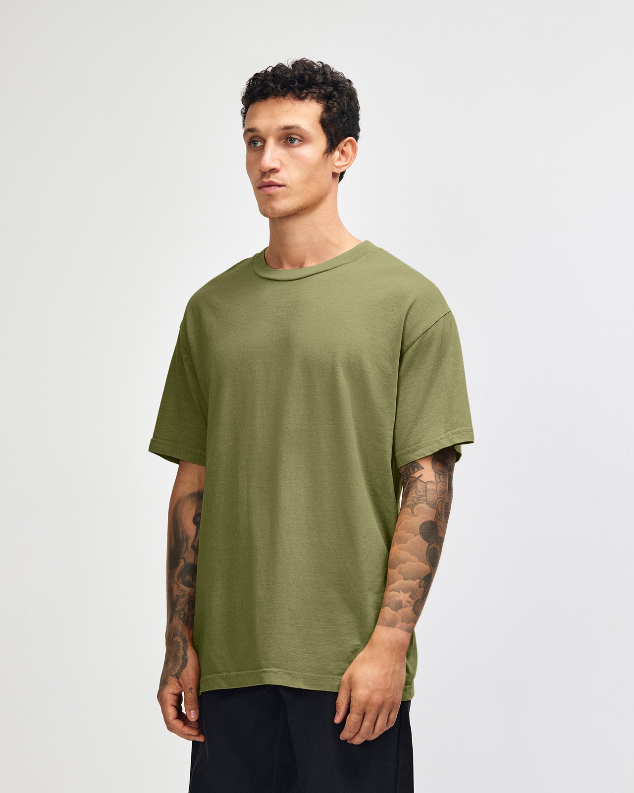 Male model wearing an American Apparel Garment Dyed Heavyweight Cotton Tee Shirt in Faded Army, paired with light blue jeans, standing against a plain background (front pose) -army