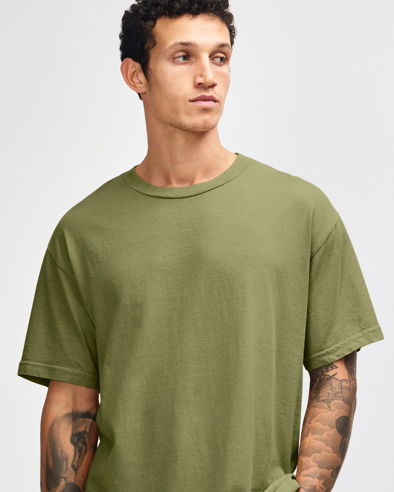 Male model wearing an American Apparel Garment Dyed Heavyweight Cotton Tee Shirt in Faded Army, paired with light blue jeans, standing against a plain background (front pose) -army
