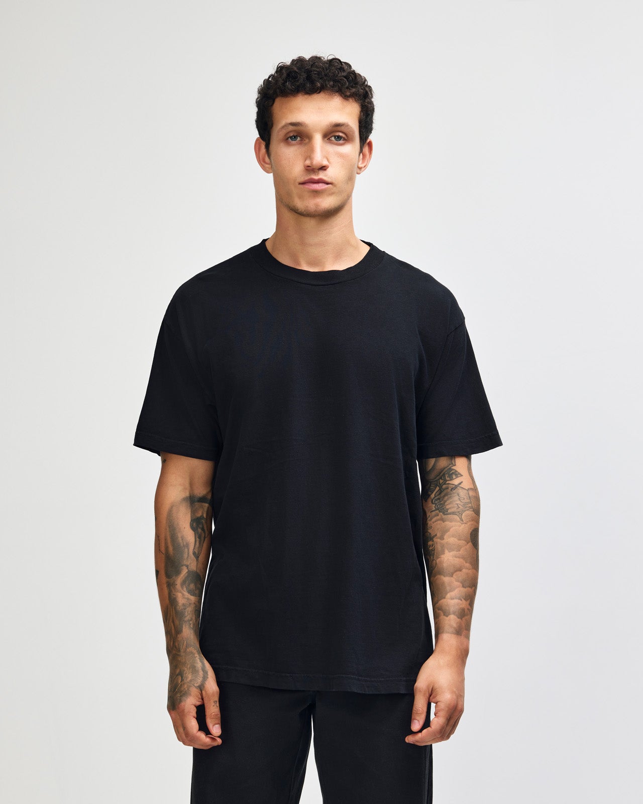 Male model wearing an American Apparel Garment Dyed Heavyweight Cotton Tee Shirt in True Black, paired with light blue jeans, standing against a plain background (front pose) -true black