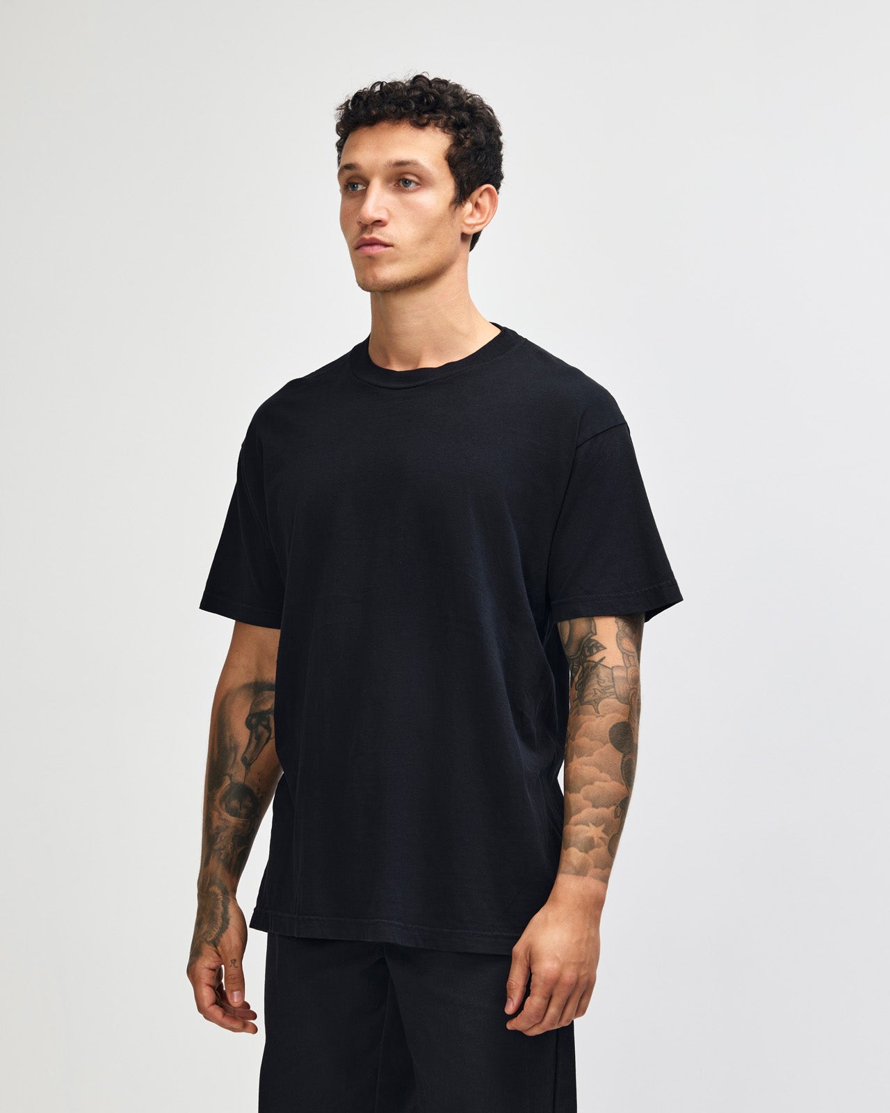 Male model wearing an American Apparel Garment Dyed Heavyweight Cotton Tee Shirt in True Black, paired with light blue jeans, standing against a plain background (front pose) -true black