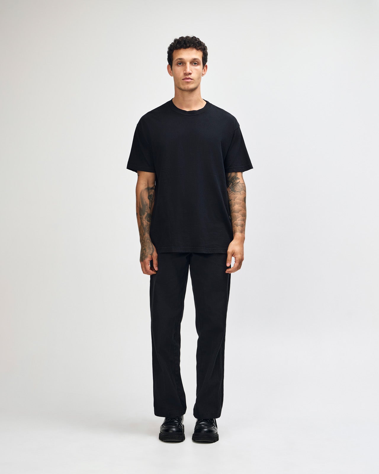 Male model wearing an American Apparel Garment Dyed Heavyweight Cotton Tee Shirt in True Black, paired with light blue jeans, standing against a plain background (front pose) -true black