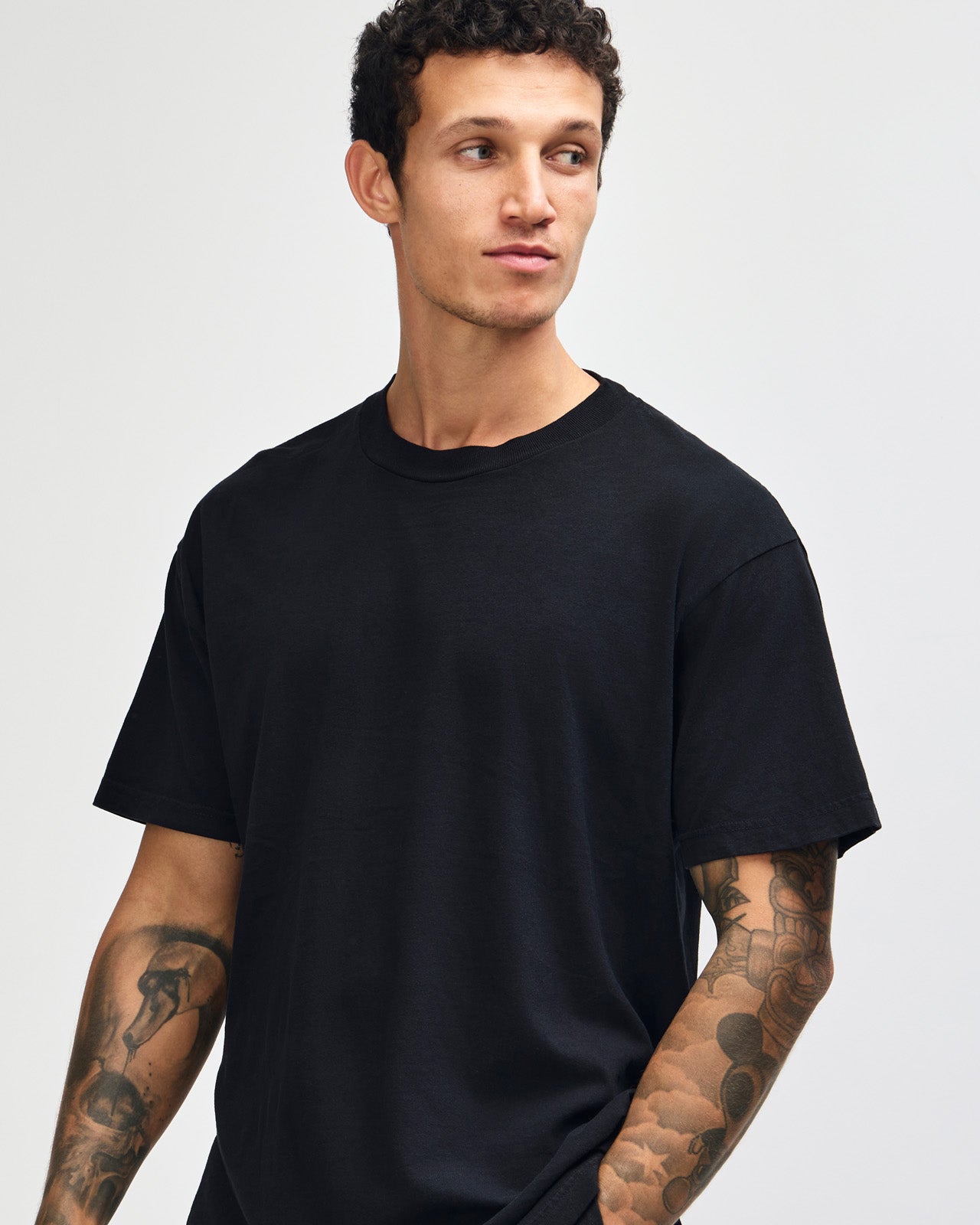Male model wearing an American Apparel Garment Dyed Heavyweight Cotton Tee Shirt in True Black, paired with light blue jeans, standing against a plain background (front pose) -true black