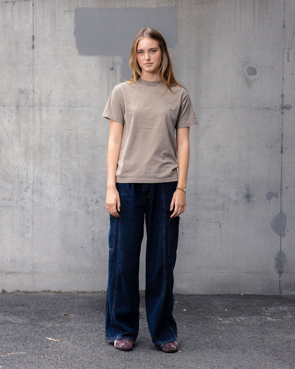 Female model wearing an American Apparel Garment Dyed Heavyweight Cotton Tee Shirt in Faded Brown, paired with light blue jeans, standing against a plain background (front pose) -faded brown