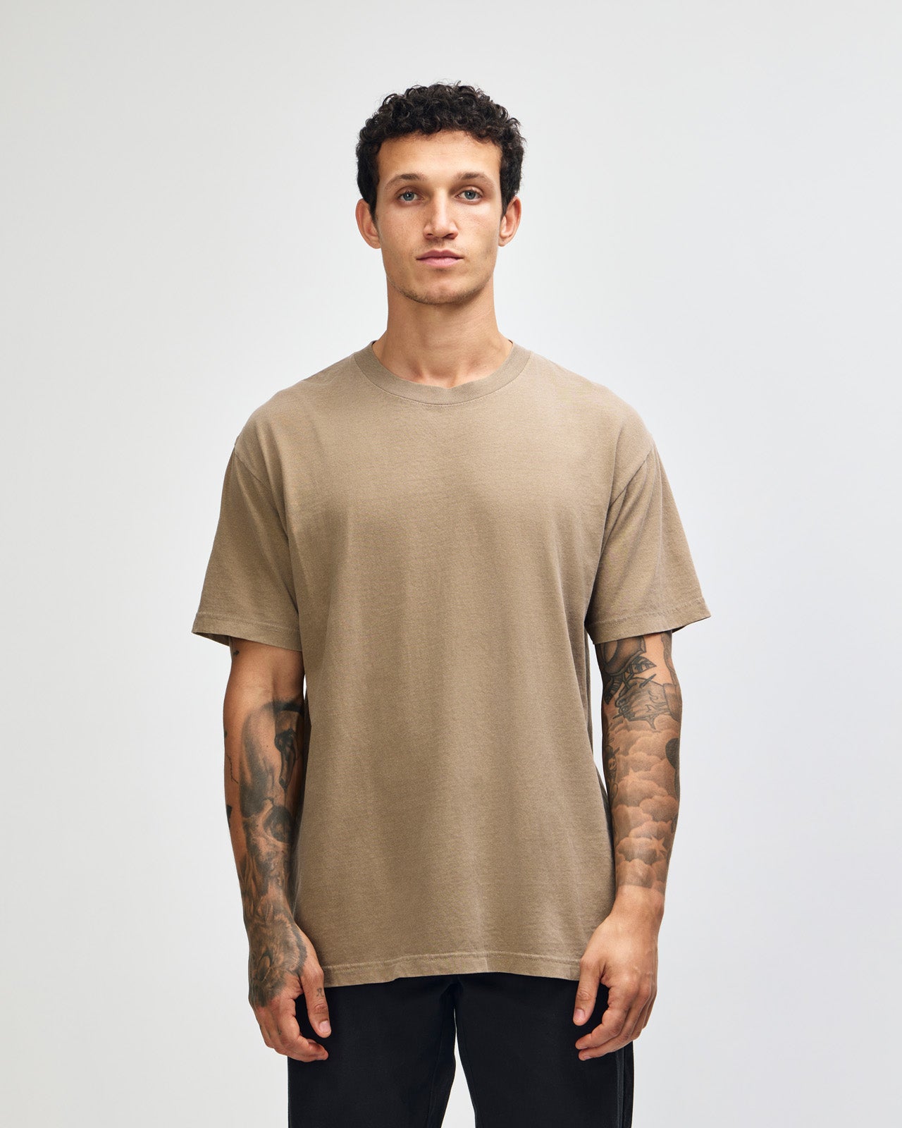 Male model wearing an American Apparel Garment Dyed Heavyweight Cotton Tee Shirt in Faded Brown, paired with light blue jeans, standing against a plain background (front pose) -brown
