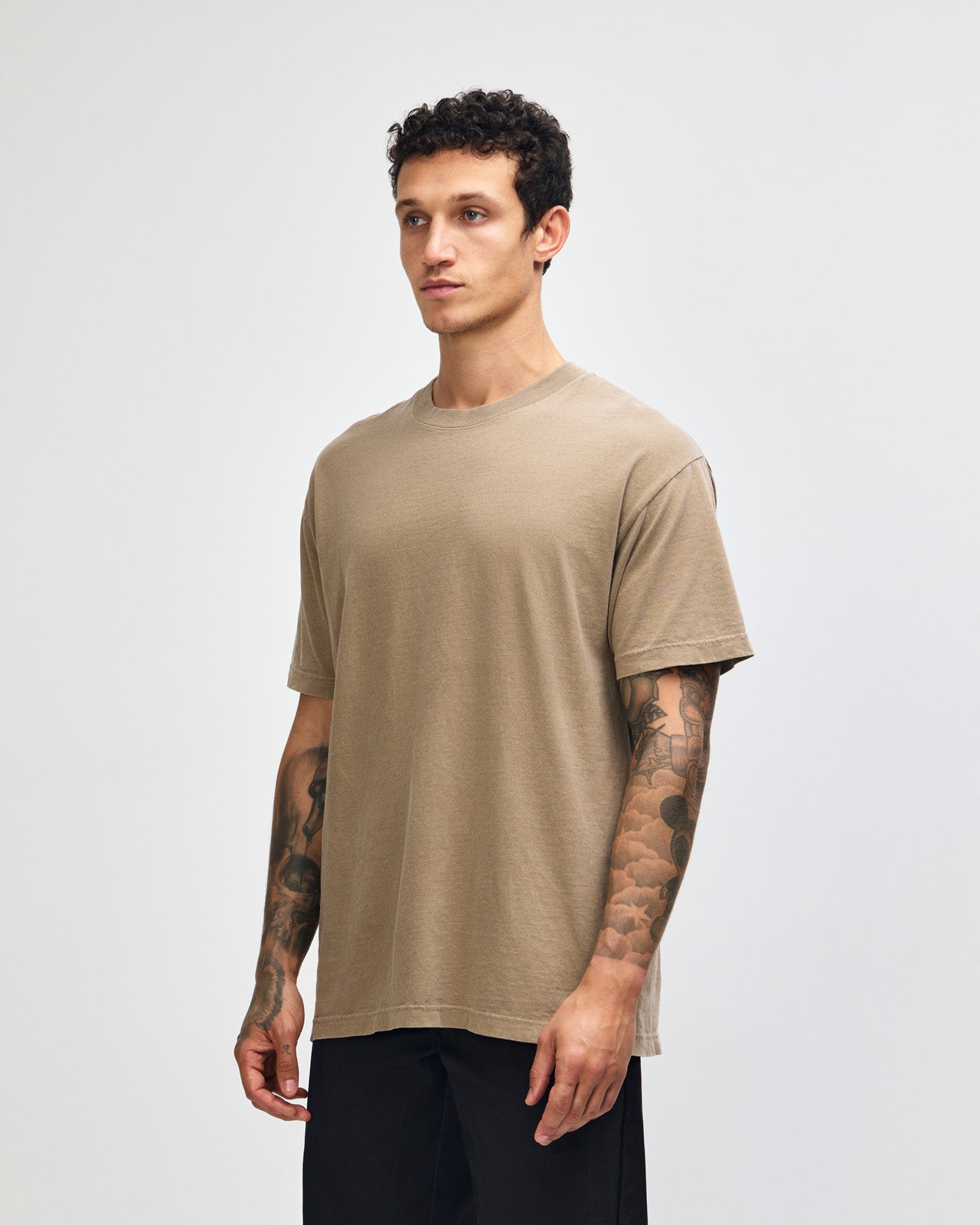 Male model wearing an American Apparel Garment Dyed Heavyweight Cotton Tee Shirt in Faded Brown, paired with light blue jeans, standing against a plain background (front pose) -brown