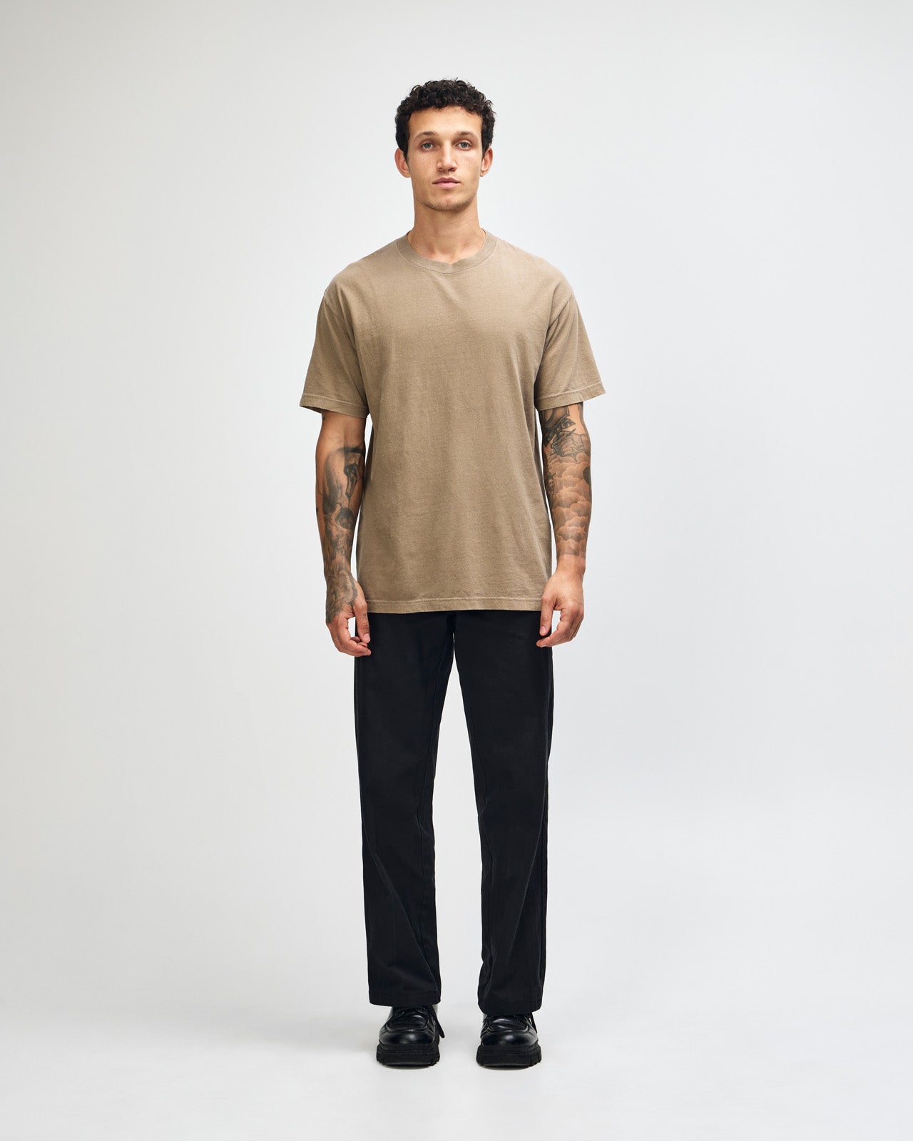 Male model wearing an American Apparel Garment Dyed Heavyweight Cotton Tee Shirt in Faded Brown, paired with light blue jeans, standing against a plain background (front pose) -brown