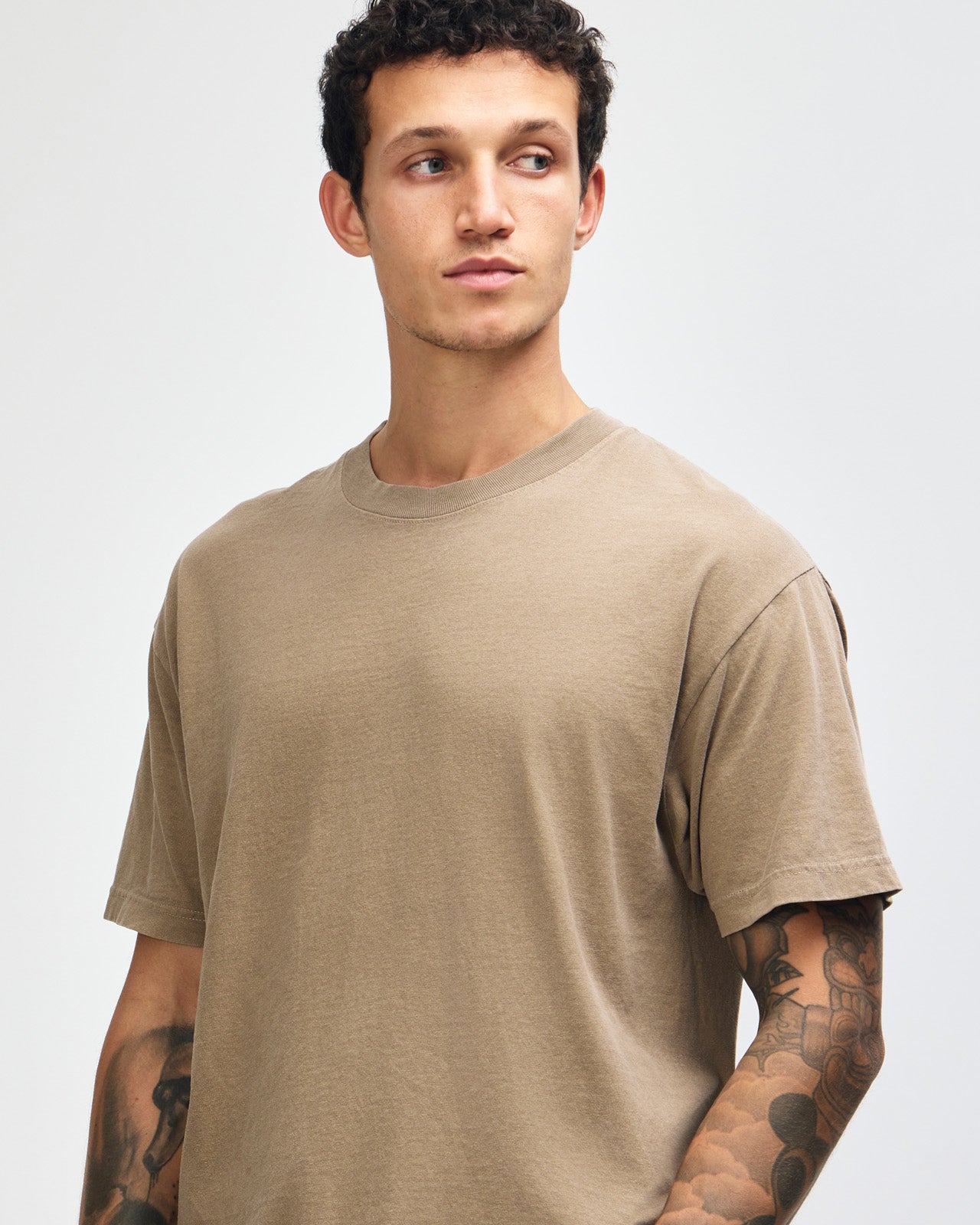 Male model wearing an American Apparel Garment Dyed Heavyweight Cotton Tee Shirt in Faded Brown, paired with light blue jeans, standing against a plain background (front pose) -brown