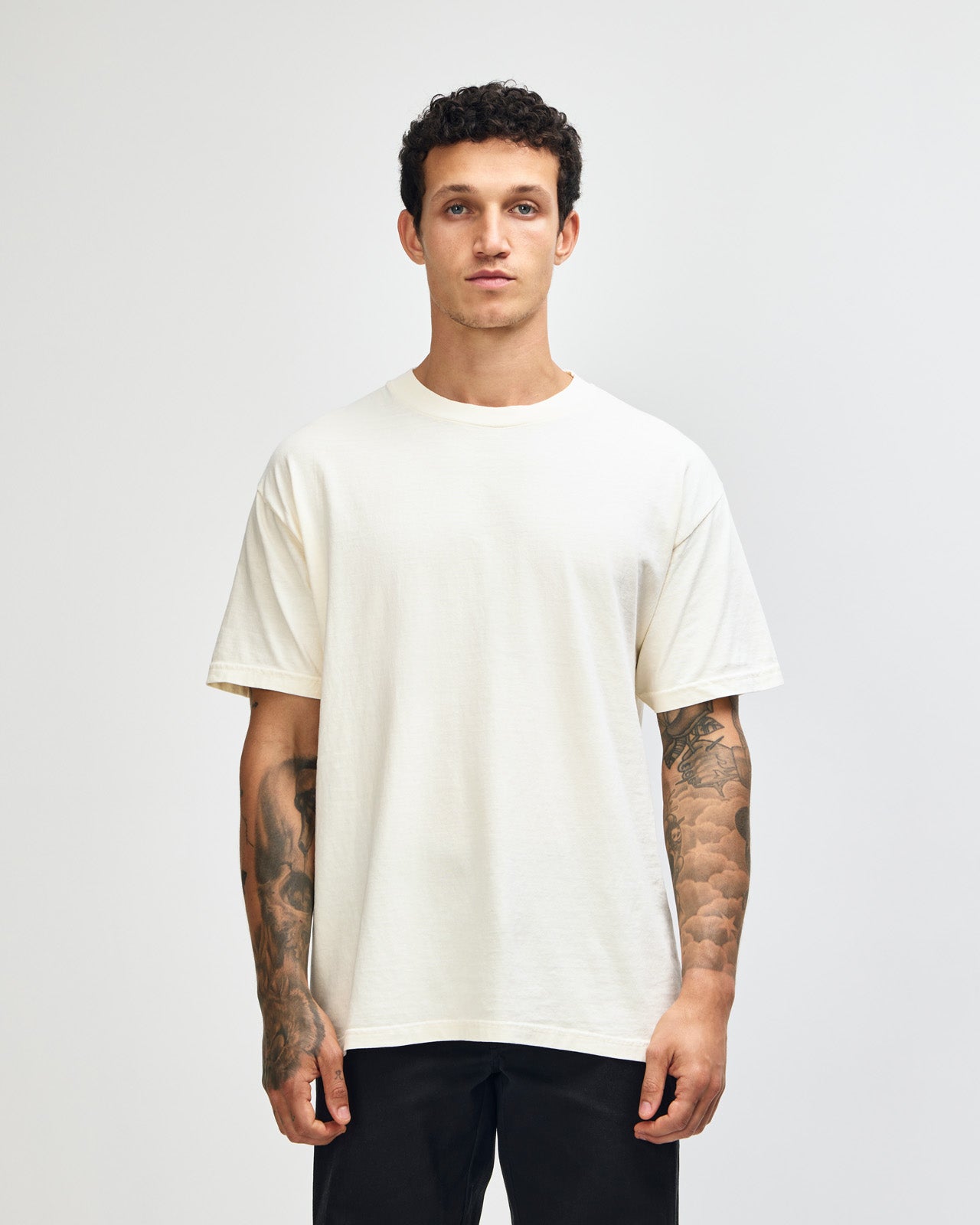 Male model wearing an American Apparel Garment Dyed Heavyweight Cotton Tee Shirt in Faded Cream, paired with light blue jeans, standing against a plain background (front pose) -cream