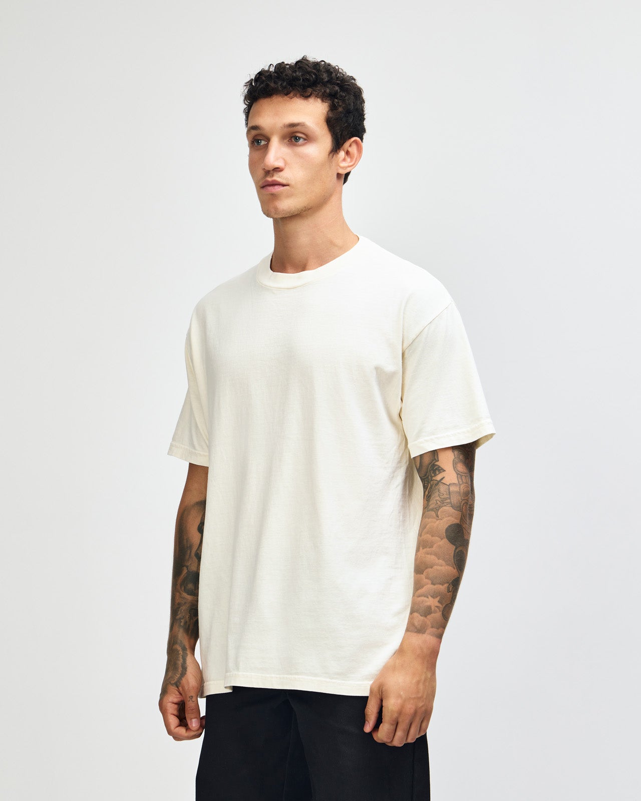 Male model wearing an American Apparel Garment Dyed Heavyweight Cotton Tee Shirt in Faded Cream, paired with light blue jeans, standing against a plain background (front pose) -cream