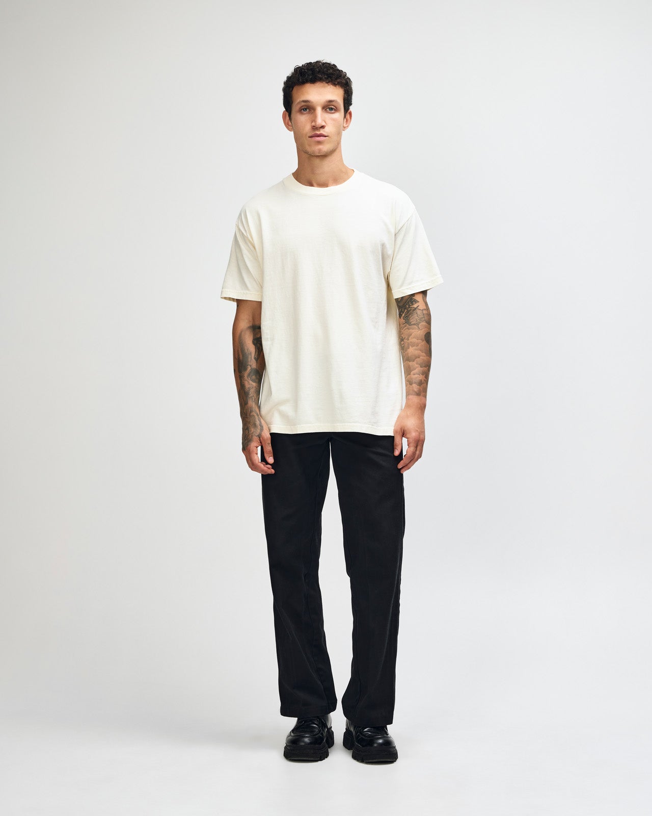 Male model wearing an American Apparel Garment Dyed Heavyweight Cotton Tee Shirt in Faded Cream, paired with light blue jeans, standing against a plain background (front pose) -cream
