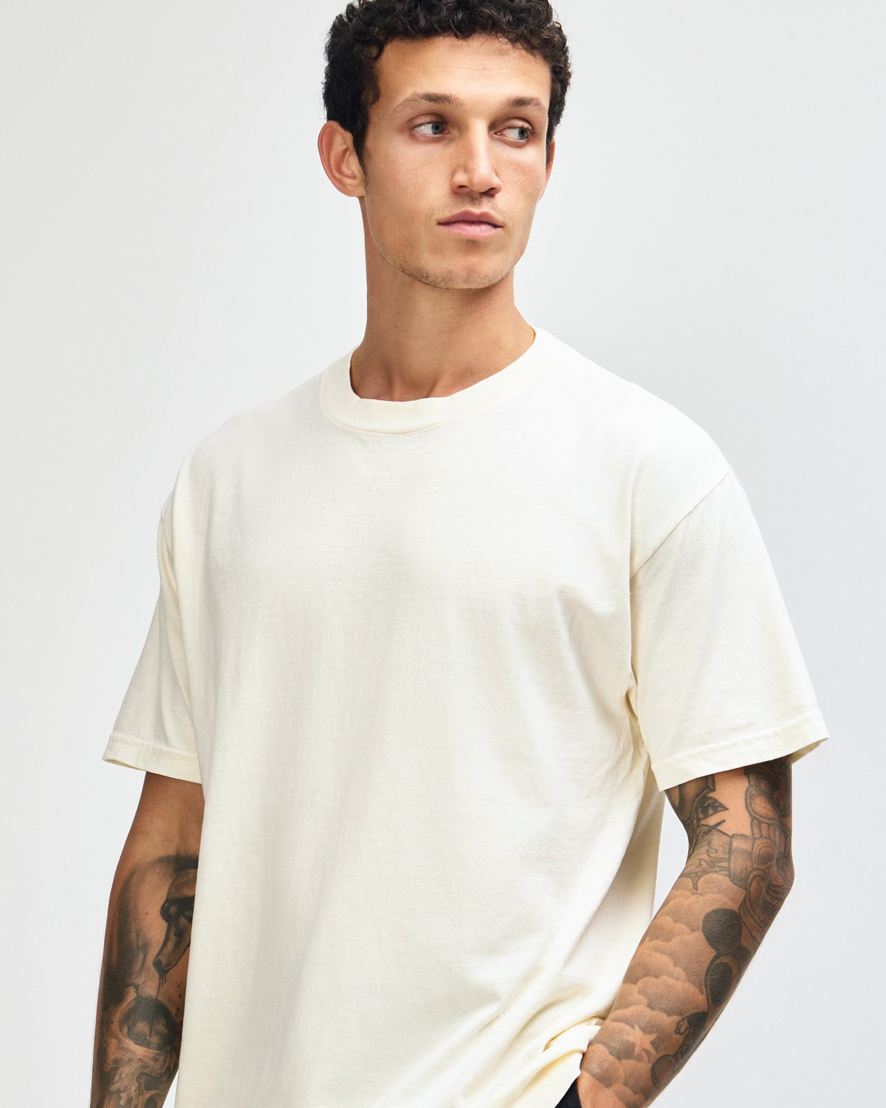 Male model wearing an American Apparel Garment Dyed Heavyweight Cotton Tee Shirt in Faded Cream, paired with light blue jeans, standing against a plain background (front pose) -cream