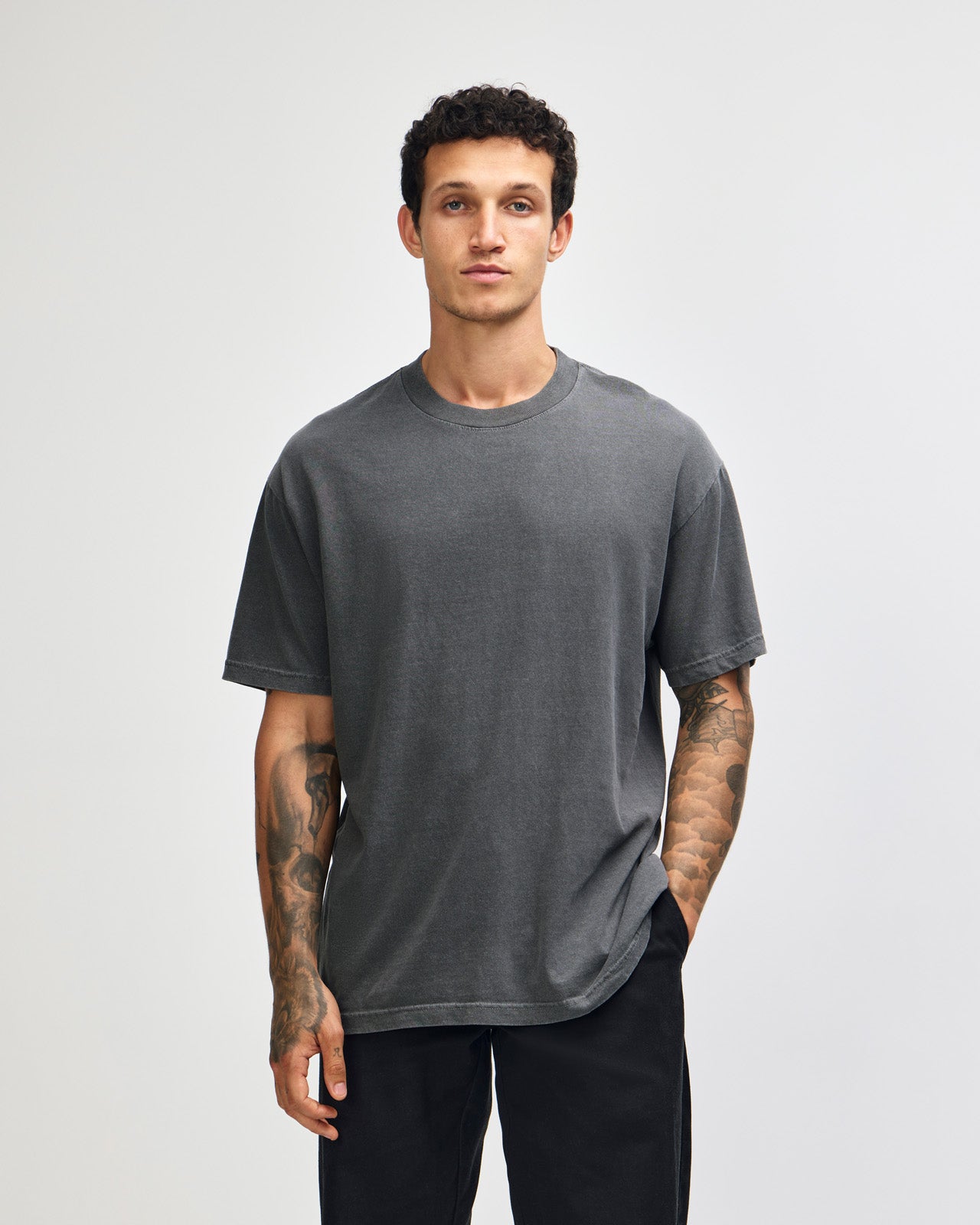 Male model wearing an American Apparel Garment Dyed Heavyweight Cotton Tee Shirt in Faded Black, paired with light blue jeans, standing against a plain background (front pose) -black