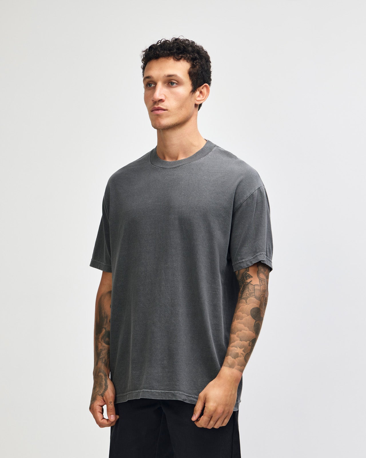 Male model wearing an American Apparel Garment Dyed Heavyweight Cotton Tee Shirt in Faded Black, paired with light blue jeans, standing against a plain background (front pose) -black