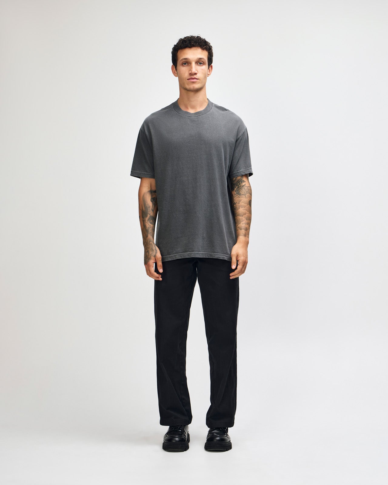 Male model wearing an American Apparel Garment Dyed Heavyweight Cotton Tee Shirt in Faded Black, paired with light blue jeans, standing against a plain background (front pose) -black