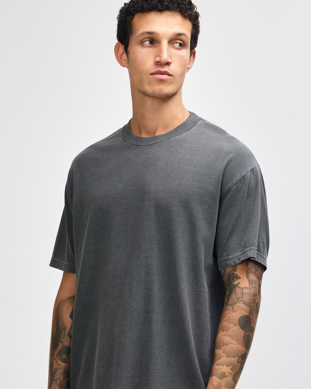 Male model wearing an American Apparel Garment Dyed Heavyweight Cotton Tee Shirt in Faded Black, paired with light blue jeans, standing against a plain background (front pose) -black