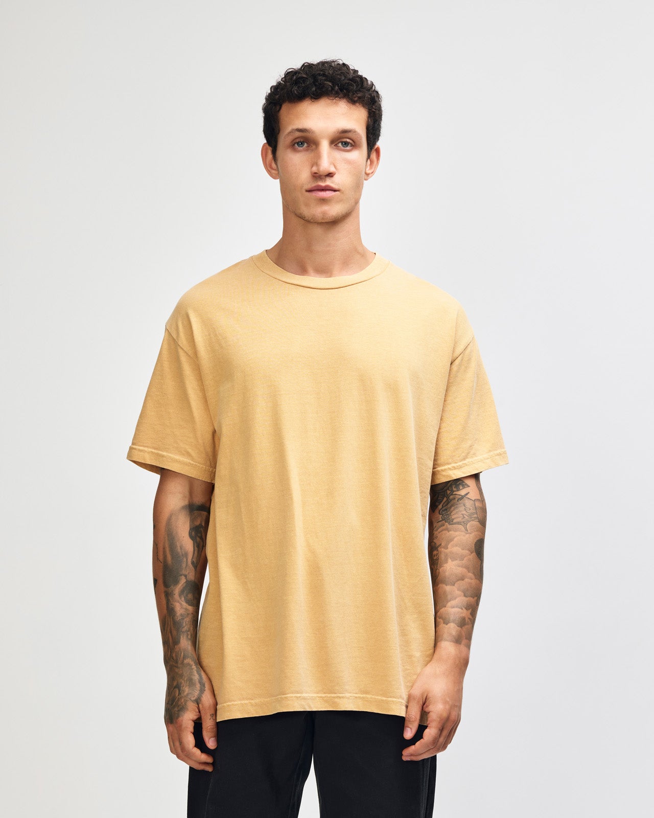 Female model wearing an American Apparel Garment Dyed Heavyweight Cotton Tee Shirt in Faded Mustard, paired with light blue jeans, standing against a plain background (front pose) -mustard