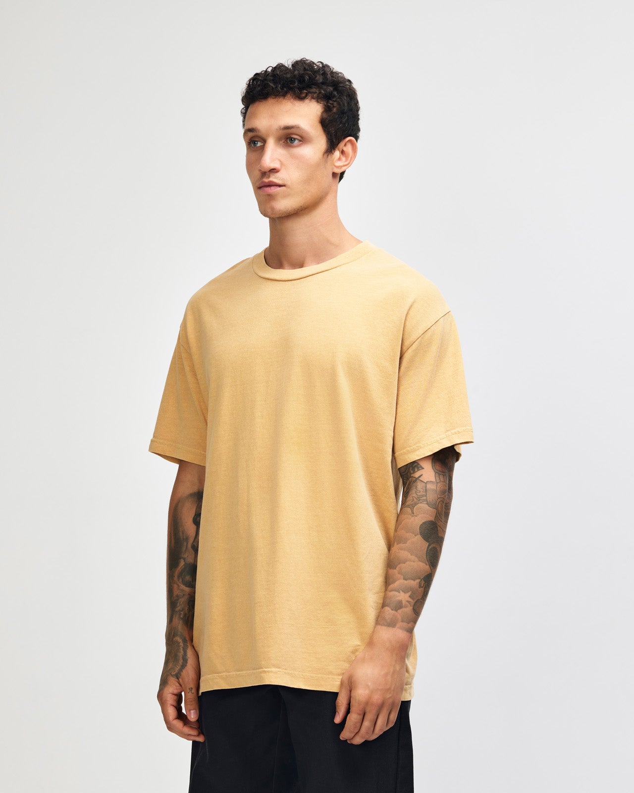 Female model wearing an American Apparel Garment Dyed Heavyweight Cotton Tee Shirt in Faded Mustard, paired with light blue jeans, standing against a plain background (front pose) -mustard