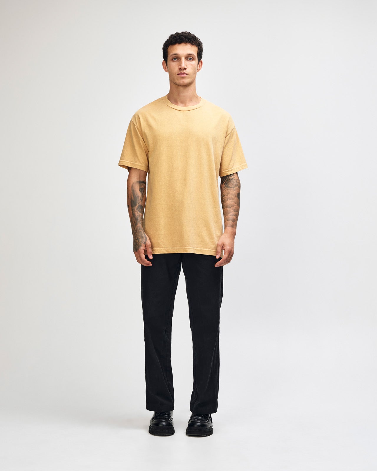 Female model wearing an American Apparel Garment Dyed Heavyweight Cotton Tee Shirt in Faded Mustard, paired with light blue jeans, standing against a plain background (front pose) -mustard