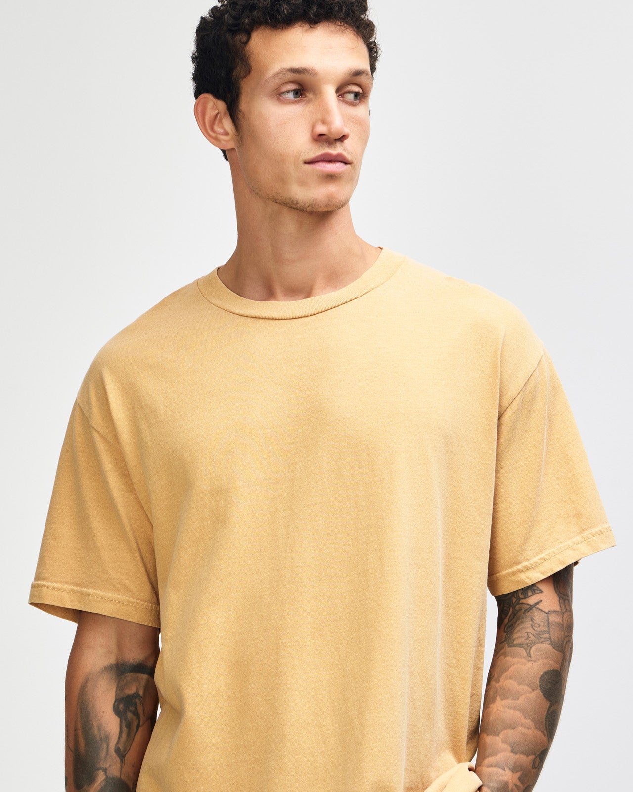 Female model wearing an American Apparel Garment Dyed Heavyweight Cotton Tee Shirt in Faded Mustard, paired with light blue jeans, standing against a plain background (front pose) -mustard