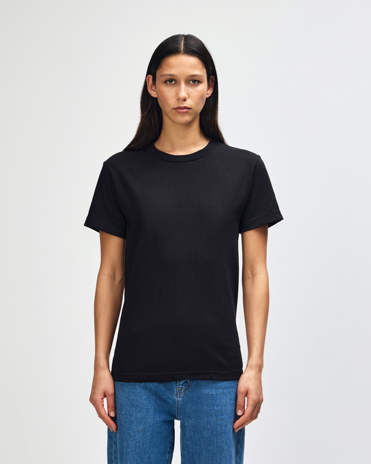 Female Model wearing a black Heavyweight Unisex Short Sleeve Crew Neck Tee, paired with blue jeans, standing against a plain background (front pose) -black