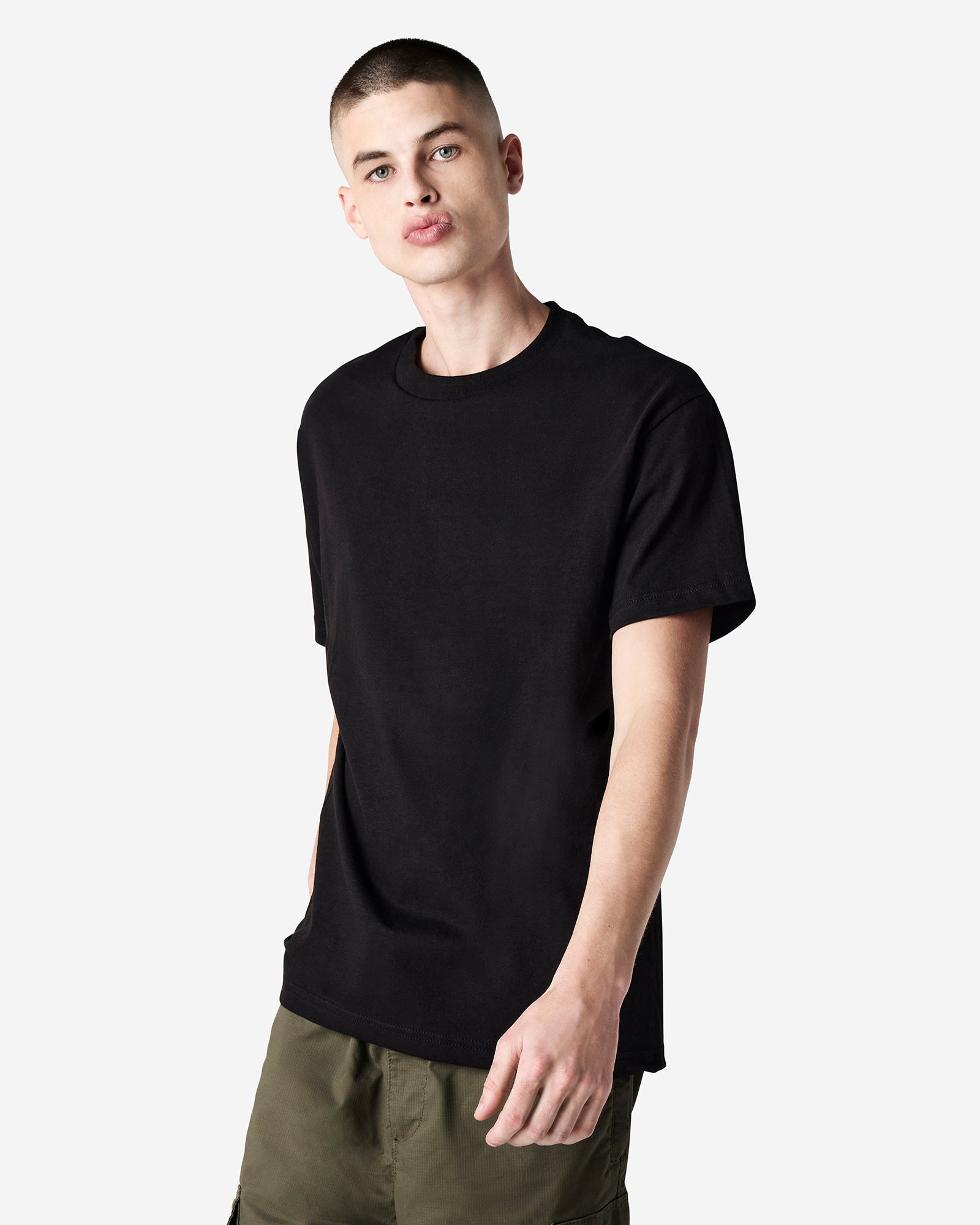 Male Model wearing a Black Heavyweight Unisex Short Sleeve Crew Neck Tee, paired with green jeans, standing against a plain background (front pose) -black