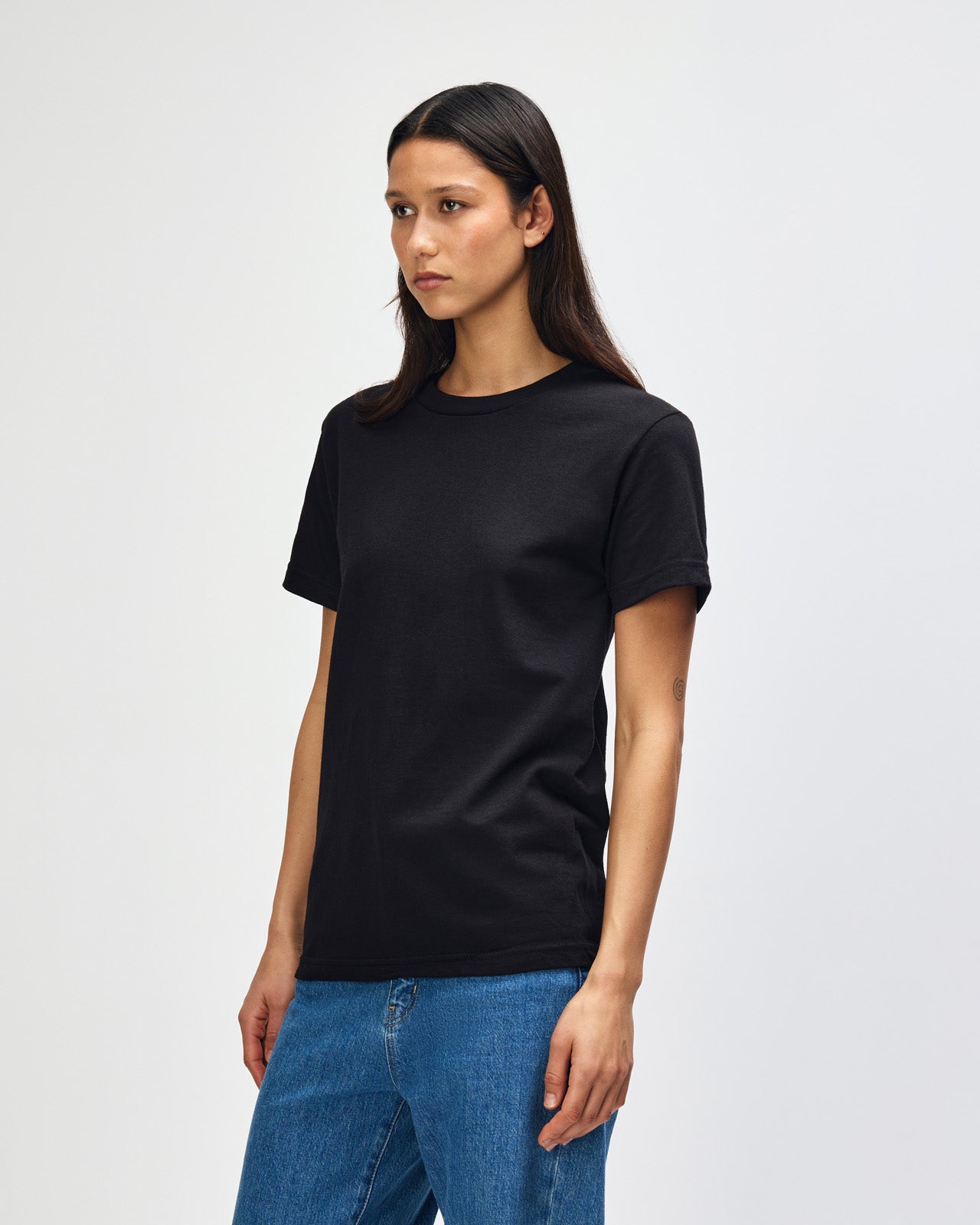 Female Model wearing a black Heavyweight Unisex Short Sleeve Crew Neck Tee, paired with blue jeans, standing against a plain background (front pose) -black