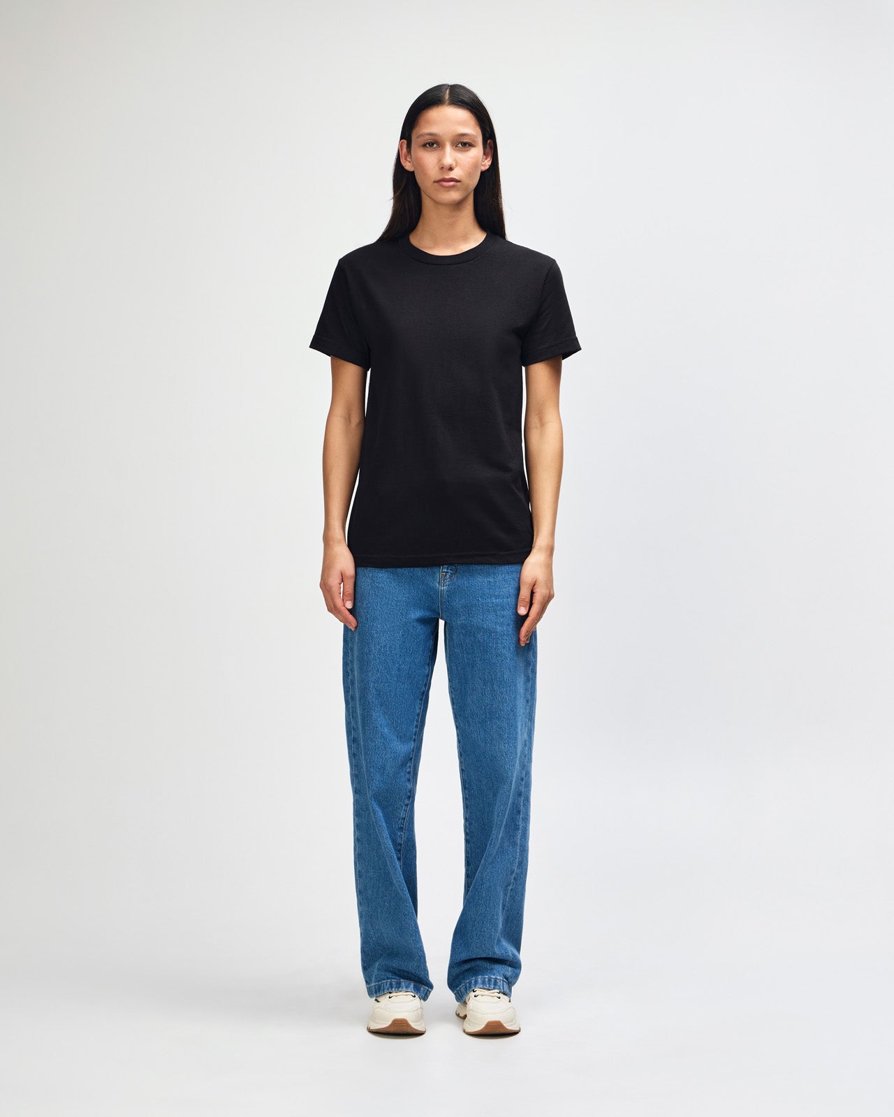 Female Model wearing a black Heavyweight Unisex Short Sleeve Crew Neck Tee, paired with blue jeans, standing against a plain background (front pose) -black