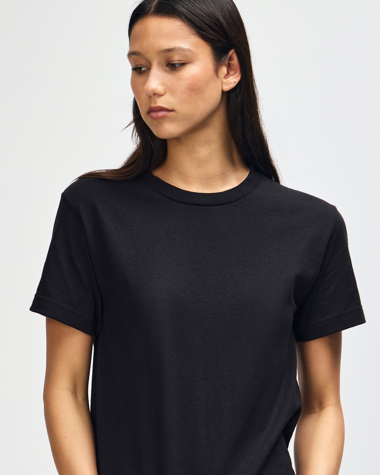 Female Model wearing a black Heavyweight Unisex Short Sleeve Crew Neck Tee, paired with blue jeans, standing against a plain background (front pose) -black
