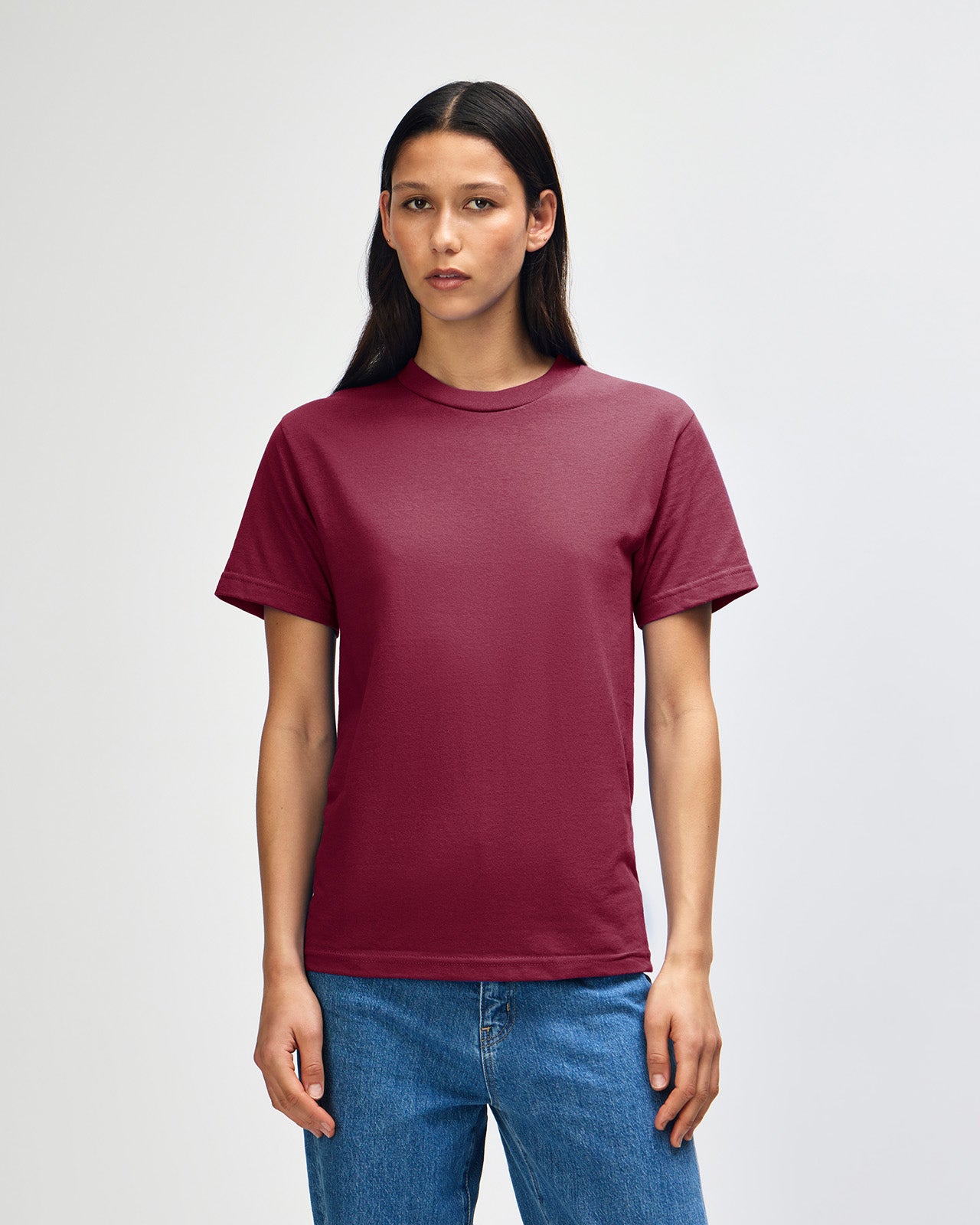 Female Model wearing a Burgundy Heavyweight Unisex Short Sleeve Crew Neck Tee, paired with blue jeans, standing against a plain background (front pose) -burgundy