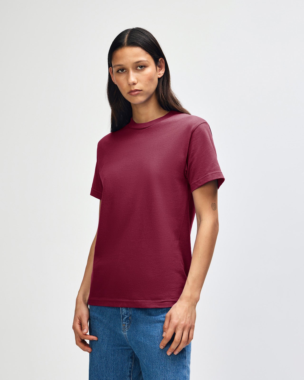 Female Model wearing a Burgundy Heavyweight Unisex Short Sleeve Crew Neck Tee, paired with blue jeans, standing against a plain background (front pose) -burgundy