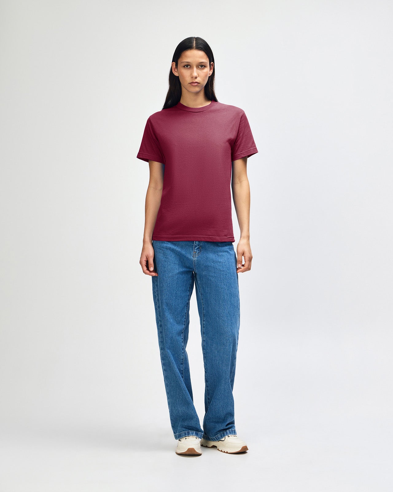 Female Model wearing a Burgundy Heavyweight Unisex Short Sleeve Crew Neck Tee, paired with blue jeans, standing against a plain background (front pose) -burgundy