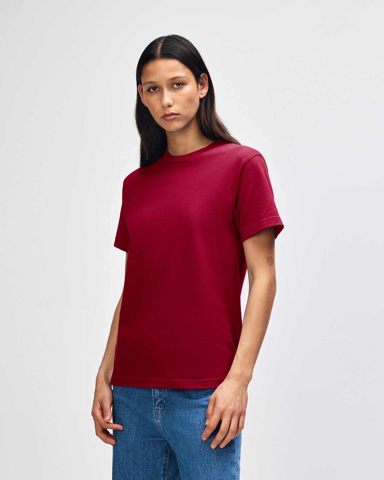 Female Model wearing a Cardinal Heavyweight Unisex Short Sleeve Crew Neck Tee, paired with blue jeans, standing against a plain background (back pose) -cardinal red 