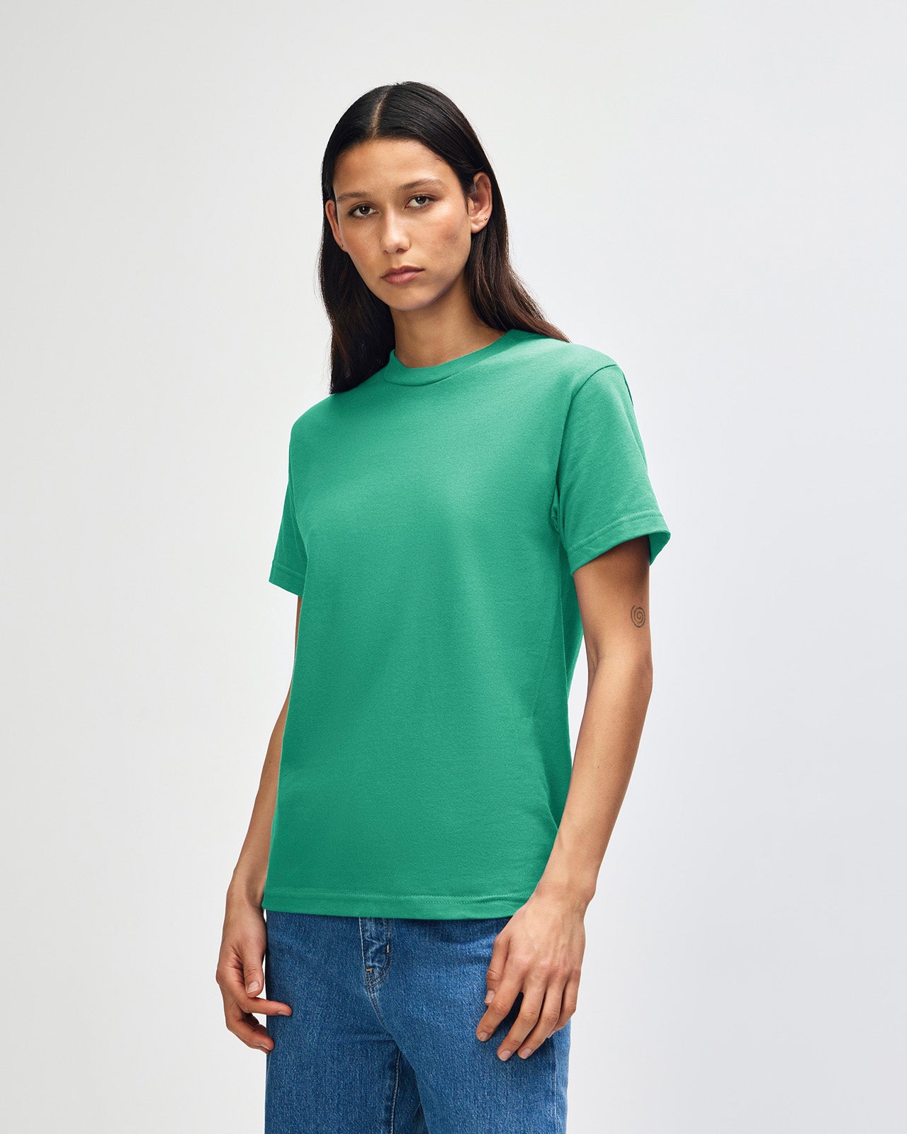 Female Model wearing a Cardinal Heavyweight Unisex Short Sleeve Crew Neck Tee, paired with blue jeans, standing against a plain background (back pose) -celadon 