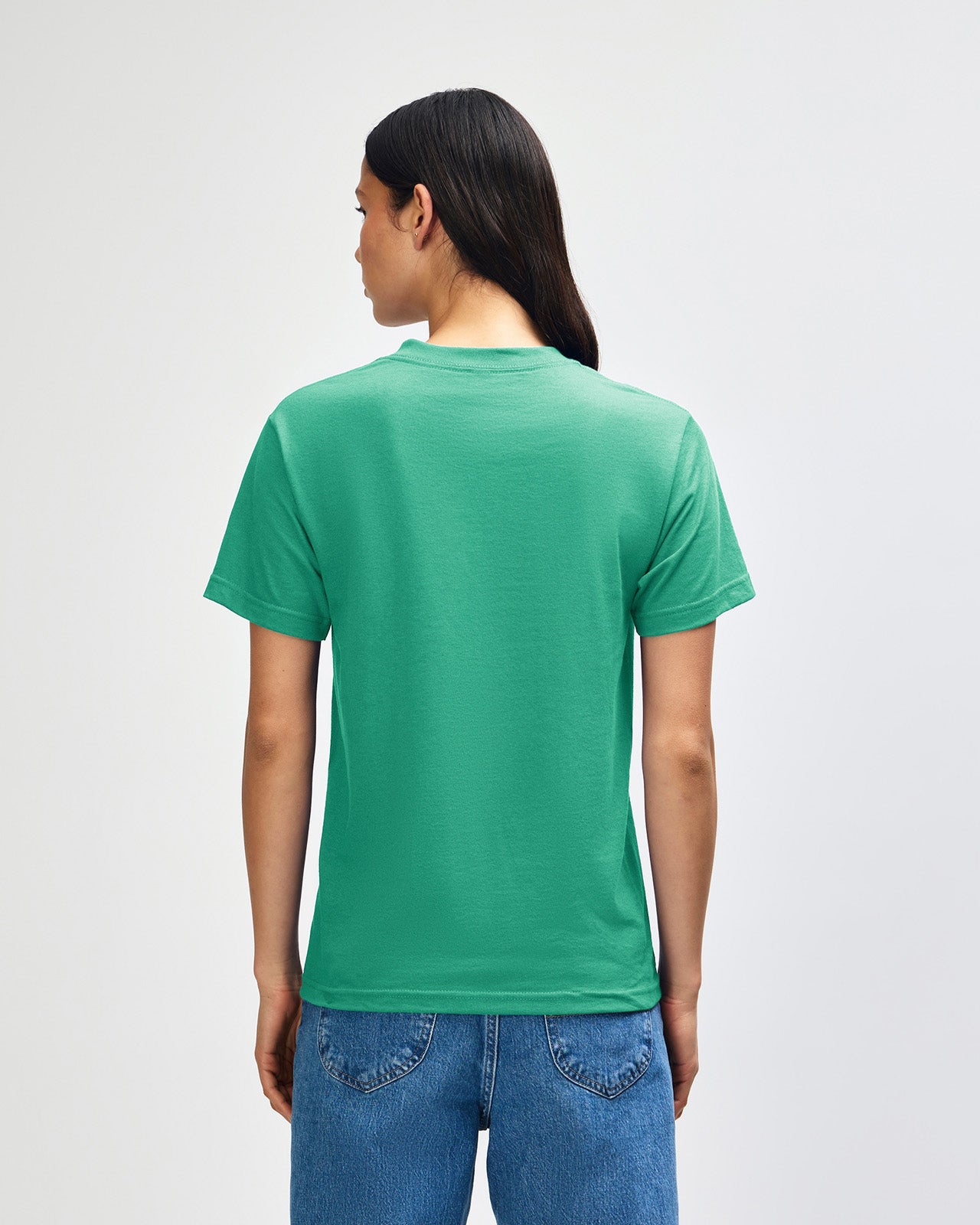Female Model wearing a Cardinal Heavyweight Unisex Short Sleeve Crew Neck Tee, paired with blue jeans, standing against a plain background (back pose) -celadon 
